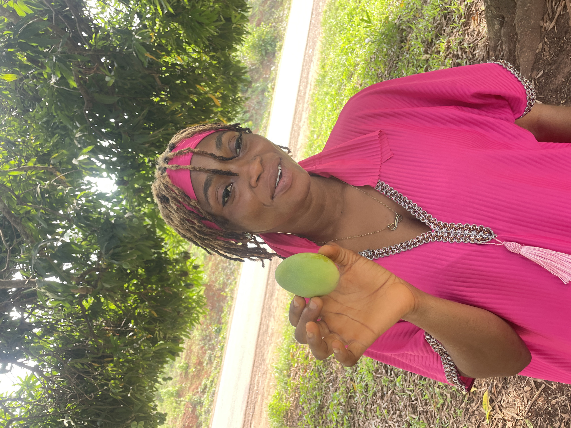 An adult woman with medium-dark skin is featured in an outdoor setting in Ogbe, Nigeria. She smiles pleasantly at the camera while holding a small, green, unripe mango in her left hand, presenting it to the viewer. Her dreadlocks are pulled back by a pink headband, which matches her vibrant pink, long-sleeved garment. The garment has a pleated texture and is adorned with intricate silver and bronze braided trim along the neckline and cuffs, complete with a pink tassel near the collar. A delicate gold chain necklace with an infinity symbol charm rests on her chest.
The scene takes place under a dense canopy of green trees, suggesting a mango orchard or a richly vegetated area. Bright natural light filters through the leaves, indicating it is daytime. In the mid-ground, a light-colored dirt path or road runs horizontally, separating the foreground's grassy terrain from further distant foliage. The woman's relaxed smile conveys a sense of contentment in this natural, tropical environment.
