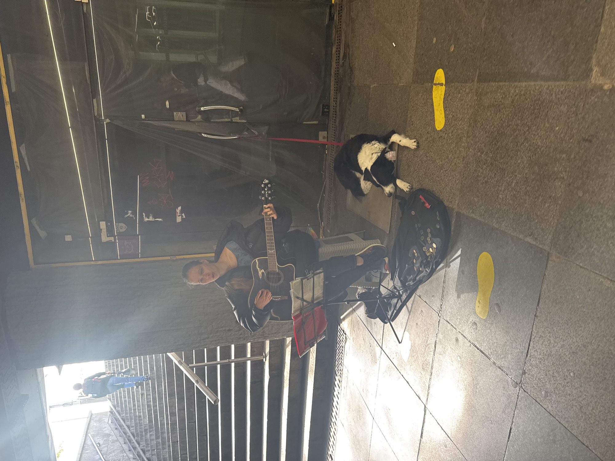 A busker sits with a black acoustic guitar and music stand, busking near stairs that lead to an underground pedestrian passageway in Sofia, Bulgaria. She is dressed in dark clothing, and the case for the guitar is nearby. A black and white dog lies on a red leash at the side of the busker. Two yellow footprints appear to be painted on the pavement. Pedestrians walk through the tunnel in the background.