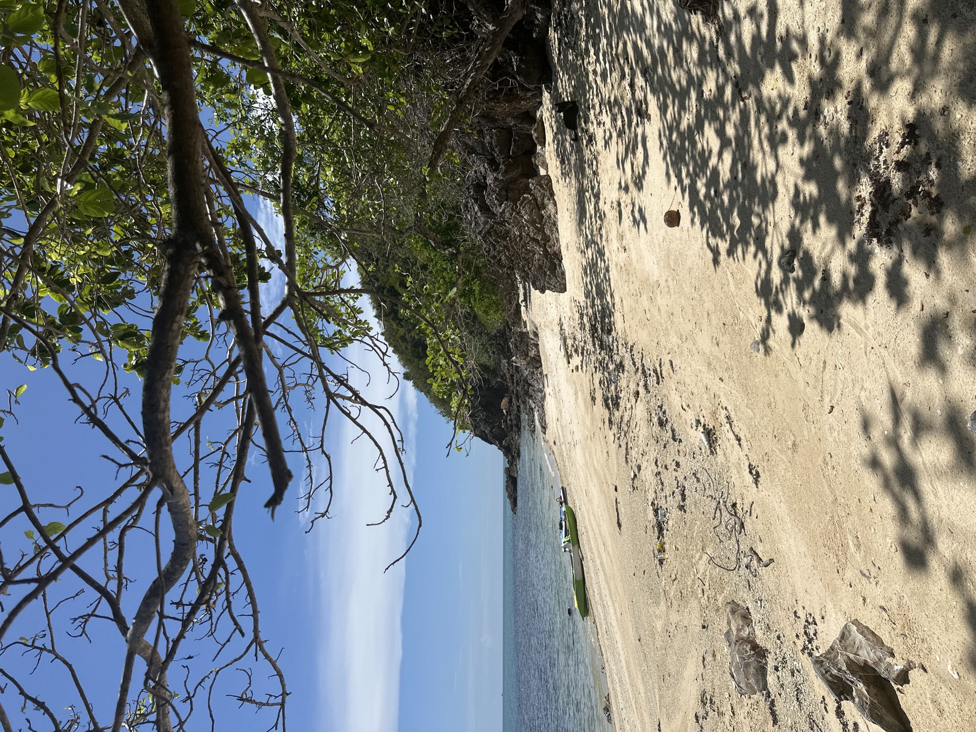 A serene beach scene is captured on the island of Koh Phangan, Thailand. The foreground is dominated by a sandy beach littered with rocks and debris, transitioning to the clear, calm ocean. Two kayaks, one green and one blue, are partially submerged near the shore. The middle ground features a rocky coastline with lush green vegetation and trees casting dappled shadows on the sand. In the distance, a hillside covered in trees slopes down towards the sea under a bright blue sky with scattered white clouds. The overall atmosphere is peaceful and natural, showcasing the tropical beauty of the location.