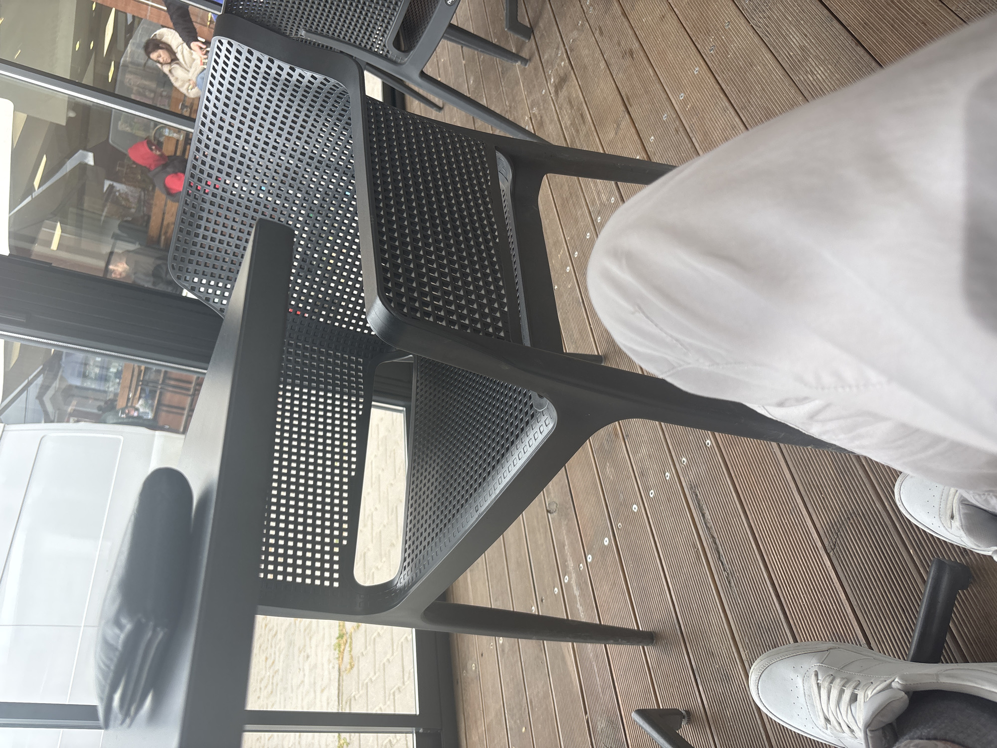 A first-person, elevated, interior shot depicts a person seated on a black, perforated plastic chair at a table on a wooden deck in Plovdiv, Bulgaria. The person's legs are visible; they are wearing white pants and white sneakers. The chairs are black, with a square-perforated design. The decking is made of narrow, evenly spaced planks running horizontally across the frame. Through a window, a blurred interior scene is visible, including other individuals. A white van can be seen through another window.