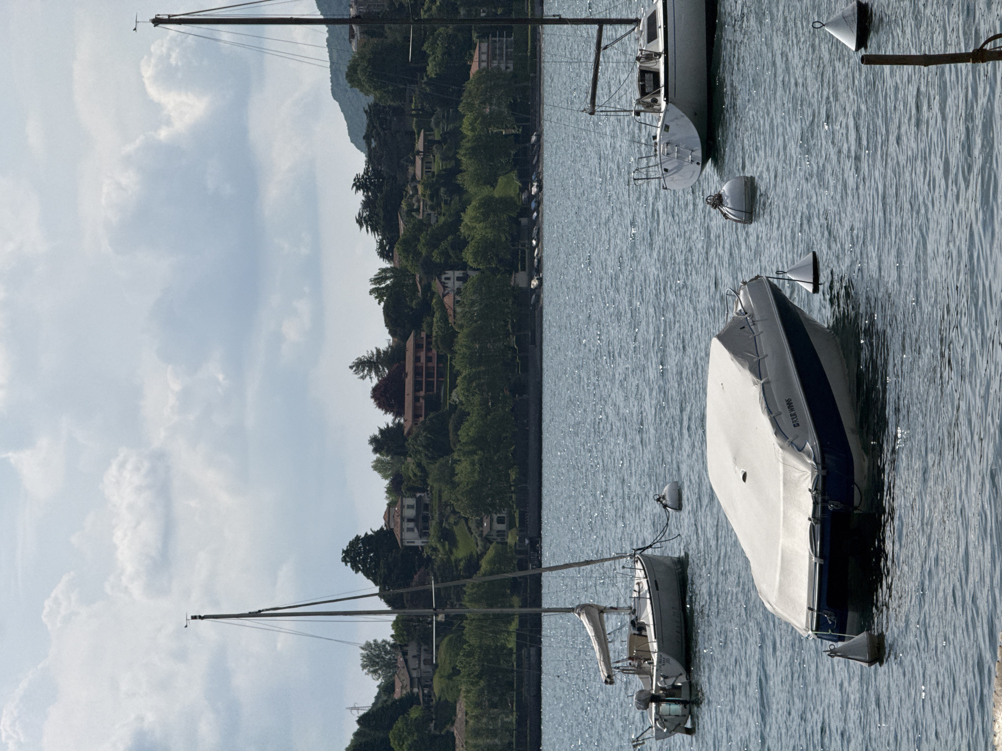 The image captures a peaceful lakeside scene, likely in Lecco, Italy, under a bright, partly cloudy sky. The foreground and midground are dominated by the calm, glistening waters of a lake, reflecting sunlight and showing gentle ripples.
Several boats are moored in the water. On the left side of the frame, a sailboat with its mast reaching high is visible, appearing to be smaller. To its right, a larger sailboat is partially seen, with its bow and part of its cabin visible, and its mast extending upwards. Further to the right, a motorboat is covered with a white protective tarp, revealing a blue hull beneath. The word "OLYMPIA" is discernible in dark lettering on the side of this covered boat. Scattered across the water are several white and black buoys.
In the background, a verdant hillside rises sharply from the water's edge. The slope is densely covered with trees, among which numerous houses and buildings are nestled, suggesting a residential area or village integrated into the natural landscape. Distant mountains are visible behind the hillside. The sky above is bright light blue with scattered white, fluffy clouds, indicating daytime and pleasant weather. The overall scene conveys a sense of tranquility and picturesque beauty.