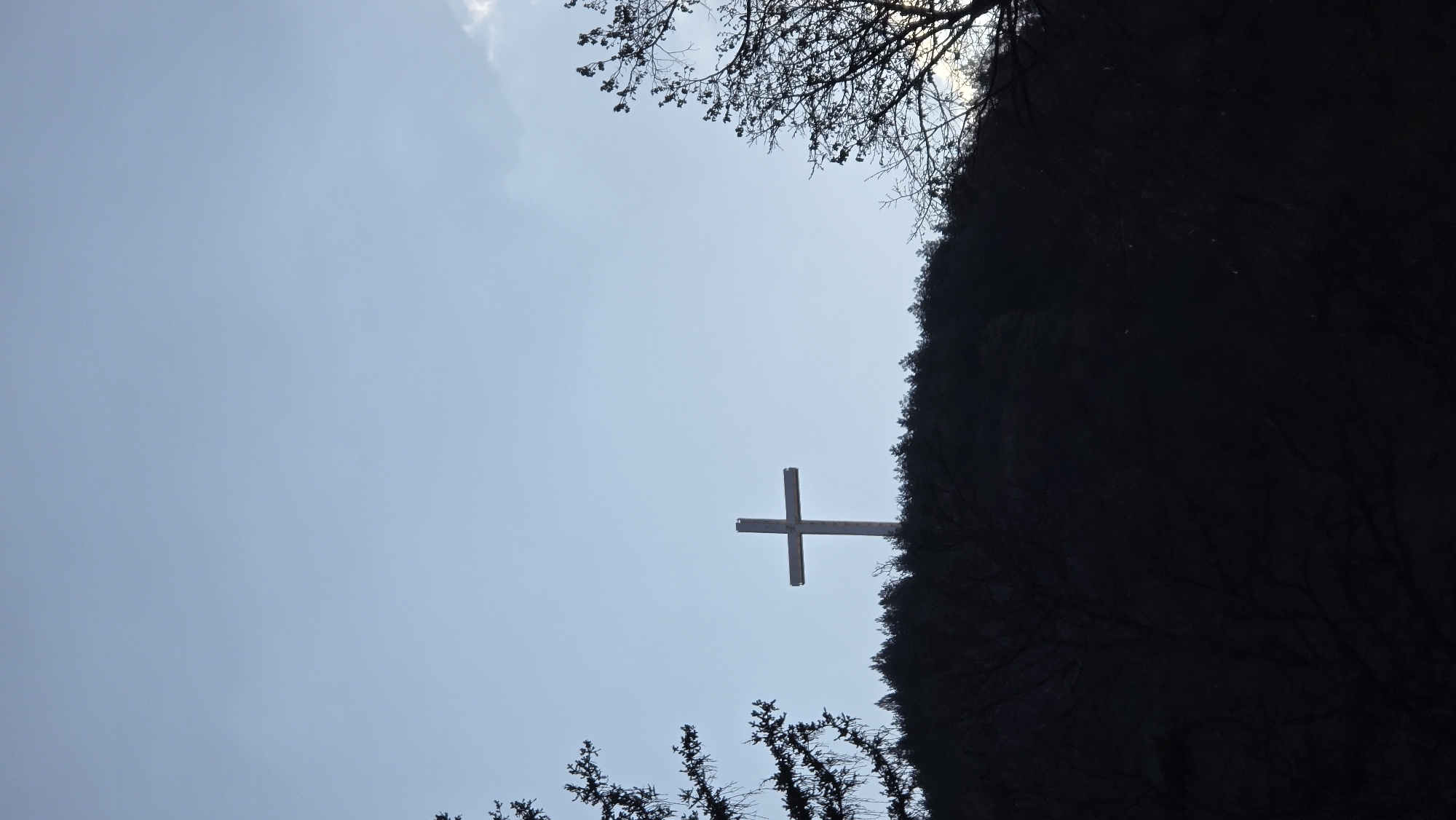 Looking up at a gray sky, a Christian cross appears to be situated on the side of a dark, tree-covered hill. Trees frame the image on top and bottom. The cross is the only distinct object in the shot. It is located in the city of Blagoevgrad, Bulgaria.