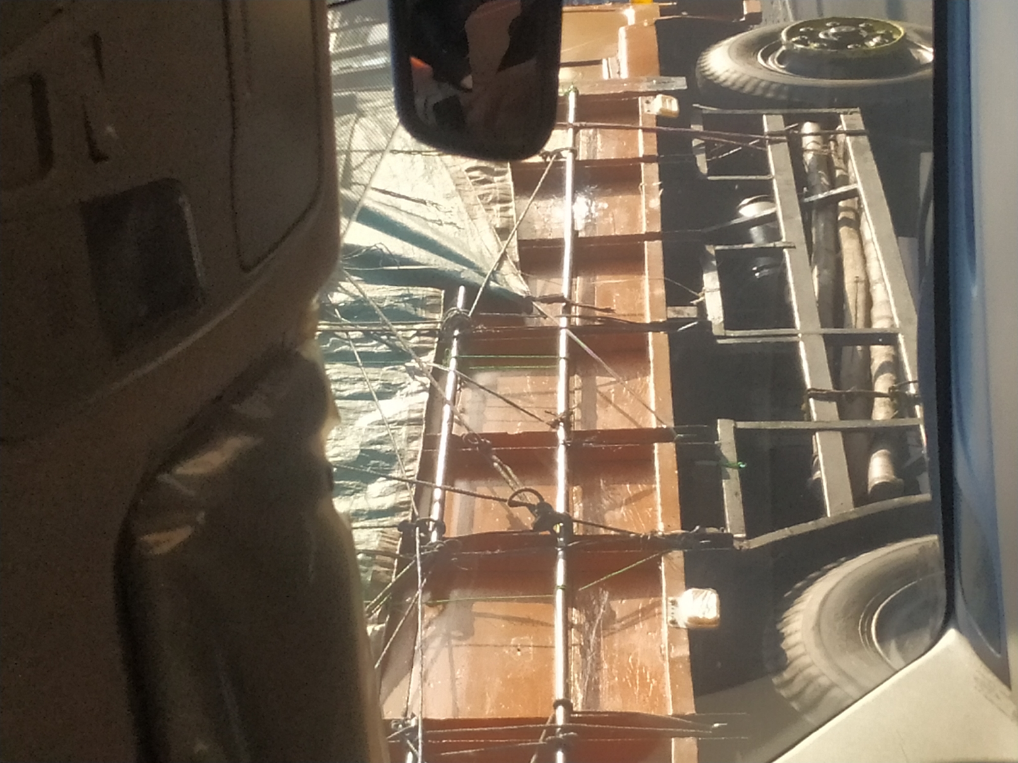 The image depicts a side view of a large, brown truck from the perspective of inside a vehicle. The truck's cargo area is covered with a light-colored tarp that is pulled taut with ropes. The side of the truck's bed appears to be made of wood or a similar material and is supported by metal framing. Various metal rods and ropes are visible, securing the tarp and the cargo. The truck has large, black tires with visible tread. A side mirror of the vehicle from which the photo is taken is visible on the right side of the image, reflecting a portion of the truck and the sky. In the foreground, the interior of the viewing vehicle is visible, including the car's ceiling liner and part of the dashboard. The environment outside the truck appears to be an outdoor setting, possibly a road, with sunlight illuminating the scene. The image is slightly grainy.