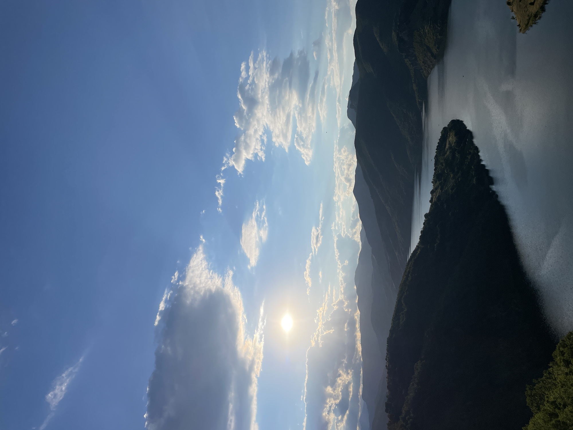 This image captures a serene landscape of rolling, tree-covered mountains embracing a tranquil, dark body of water, likely a reservoir. The sun, partially obscured by clouds, casts a warm glow across the scene, highlighting the textured cloud formations against a clear blue sky. The mountains recede into the distance, their silhouettes becoming softer and lighter, indicating depth. The water reflects the sky and surrounding greenery, creating a mirror-like effect. The location is identified as Borovitsa, Bulgaria. No people or specific actions are visible. The environment is natural and undeveloped.