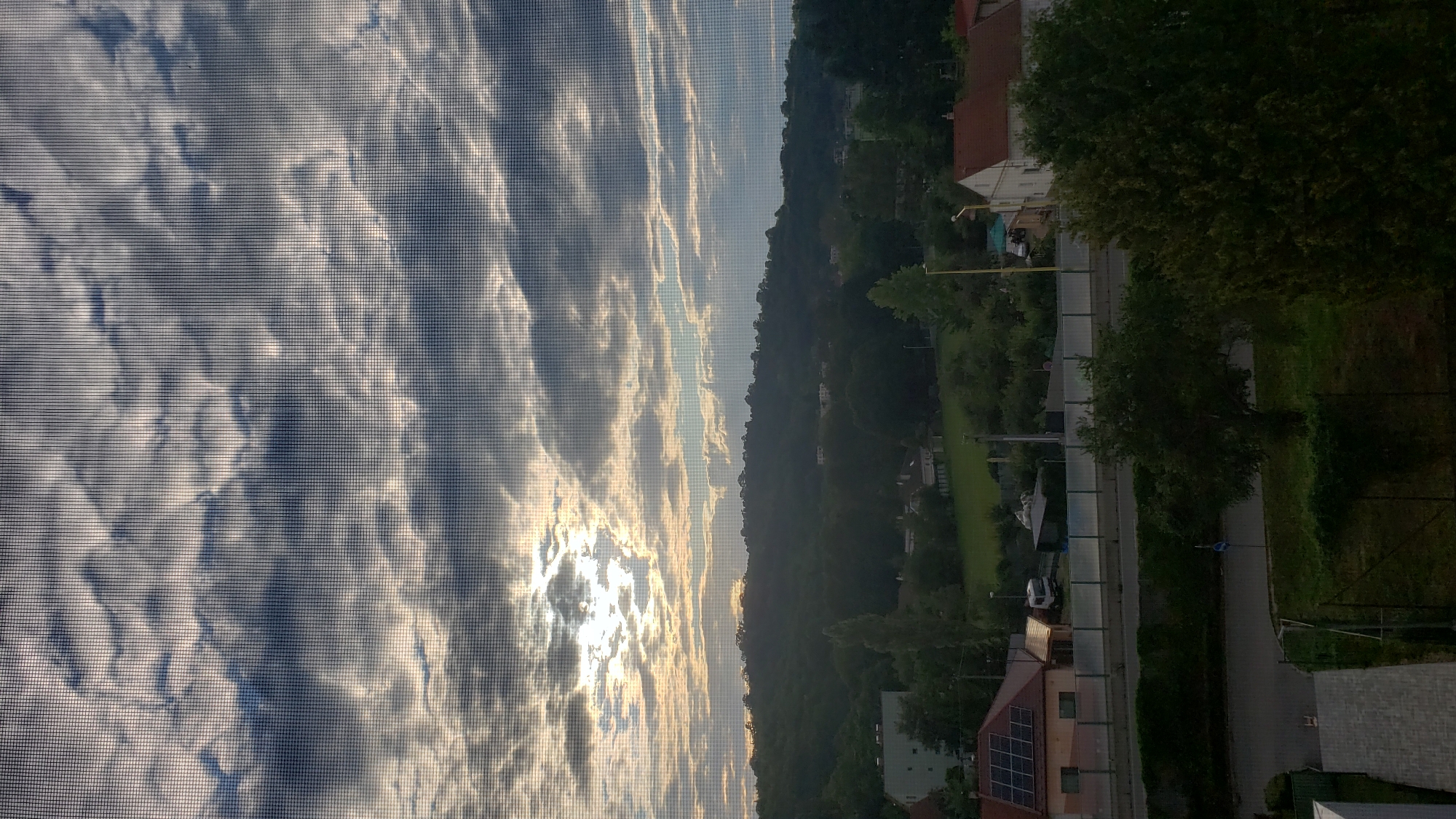 The image depicts a view of a suburban landscape through a window screen, likely captured at dawn or dusk, with the sun peeking through dramatic clouds. The foreground shows a dark, textured surface, possibly a rooftop or pavement, with a fence and some vegetation. Beyond this, a road runs horizontally, bordered by a green embankment and a sound barrier. On the other side of the road, there are houses with red-tiled roofs, solar panels on one building, and a white caravan parked nearby. A prominent green hill covered in trees rises in the background, with a few scattered houses visible on its slopes. The sky is filled with textured, layered clouds, illuminated by the low-lying sun, creating a dramatic and somewhat overcast atmosphere. The overall impression is one of a tranquil, residential area. The scene is not specifically identifiable as being within the city of Prešov, Slovakia, based solely on visual cues.