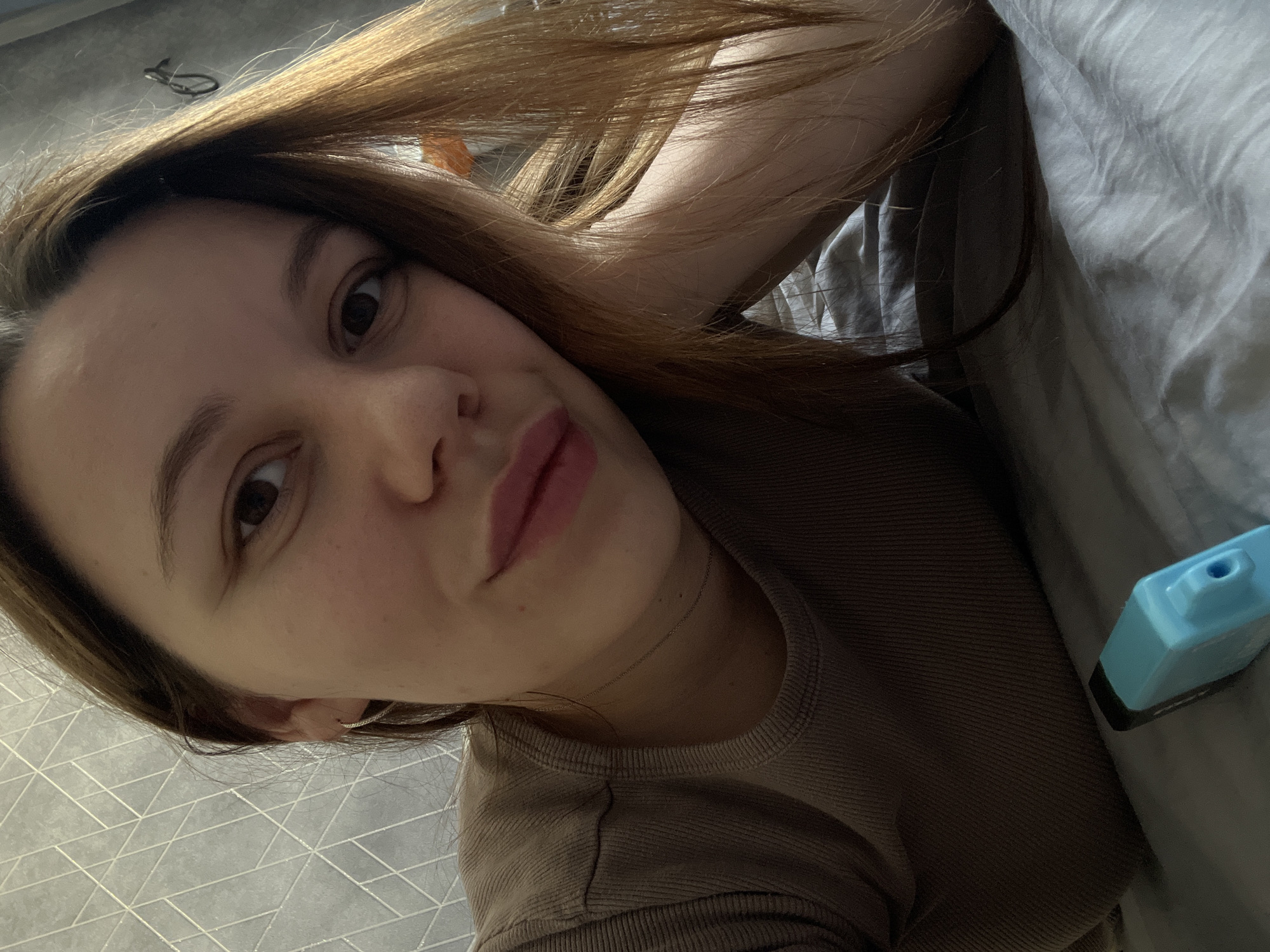 A young adult female is depicted in a close-up, head-and-shoulders selfie, observed within the city of Leninakan, Russia. She possesses light skin, dark brown eyes, and long brown hair with light brown highlights, particularly prominent on the right side of the frame. She wears a brown, ribbed, crew-neck top and a thin silver-colored chain necklace. A small hoop earring is visible in her left ear. Her lips are slightly parted in a gentle smile, displaying a light pink cosmetic application. Her head appears to be propped up by her right arm, which is mostly obscured by her hair. The background immediately behind her is a light gray wall featuring a geometric pattern of intersecting lines. To the right, a darker, plain wall section is visible with a small, dark, circular object hanging on it. She is resting on a light gray, wrinkled fabric surface, consistent with bedding or upholstery. In the lower foreground, resting on the fabric, is a light blue rectangular object with a black top and a small circular opening, consistent with a disposable vaping device.