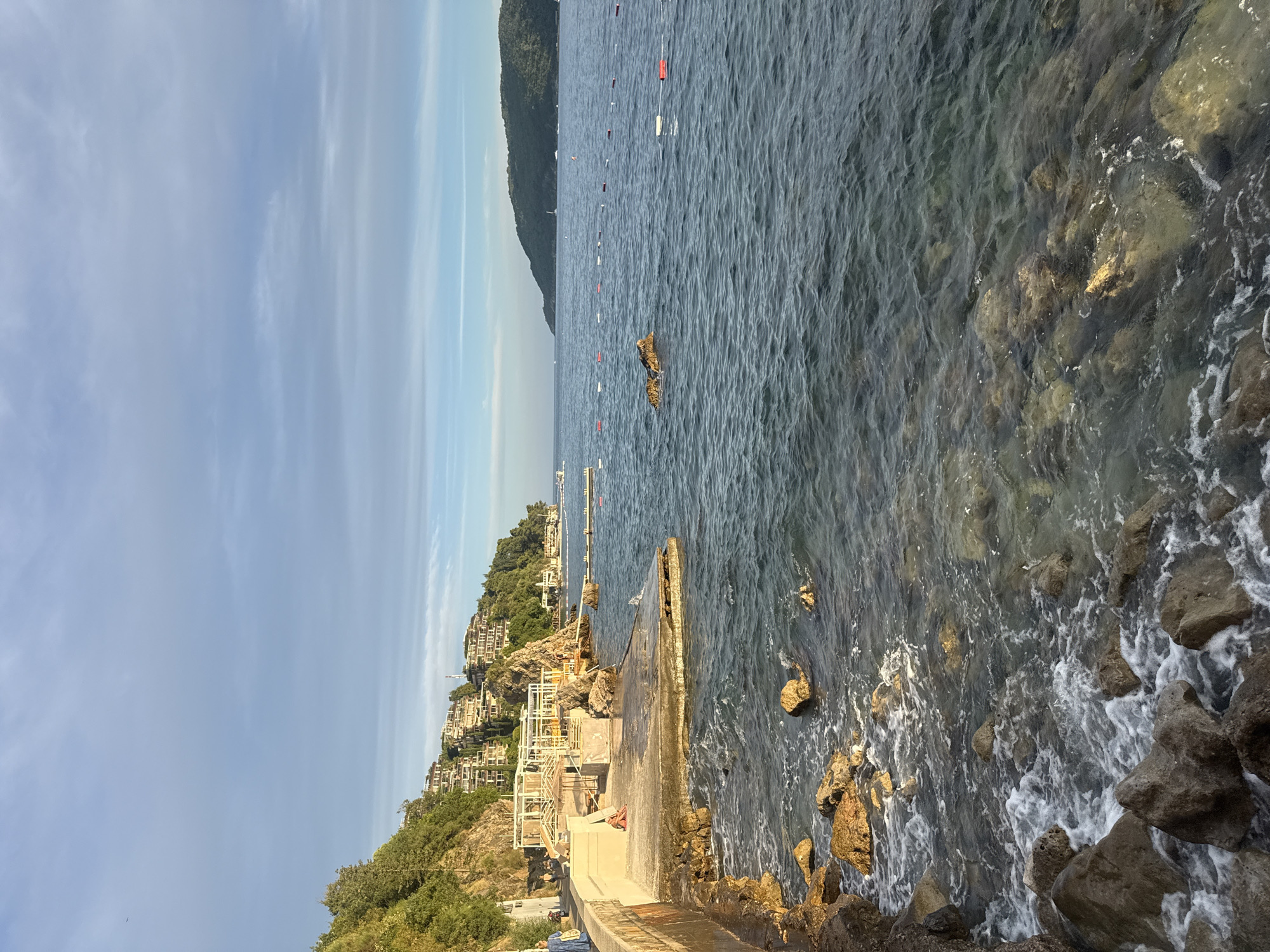 A coastal scene in Budva, Montenegro, features a rocky shoreline leading to a concrete pier and the Adriatic Sea. Buildings are visible on a lush, green hillside overlooking the water. The sky is partly cloudy with a soft, diffused light. The water is clear with small waves lapping against the rocks. Several buoys mark a swimming area in the distance. A person in a blue dress walks along the paved path on the left, and another person sits on the concrete structure.