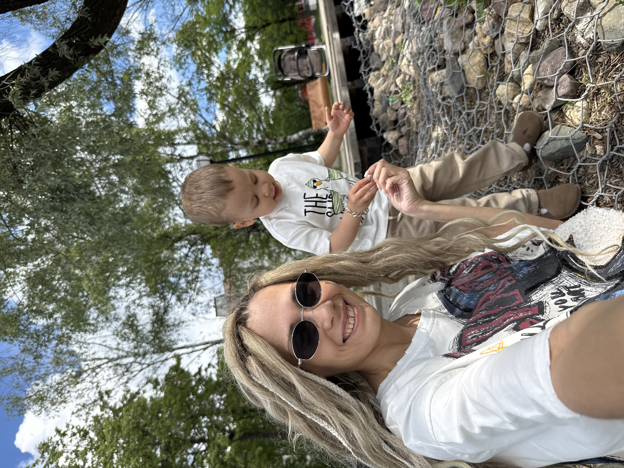 This outdoor selfie captures a smiling woman and a young child in Rusanovo, Russia, during daytime. The woman is positioned in the foreground, looking directly at the camera with a cheerful smile. She wears dark, round-framed sunglasses and a white t-shirt adorned with a large, vibrant graphic design featuring black, red, blue, and yellow brushstroke elements. Her long, light-colored hair is styled with visible braids or dreadlocks, and she wears a silver bracelet on her wrist.
Slightly behind her and to the right, a young boy with light brown hair is focused on his hands, holding a small white object. Another hand, likely the woman's, gently touches or guides his hands, indicating a shared moment of interaction or discovery. The boy is dressed in a white t-shirt that displays a cartoon graphic with the visible word "THE" followed by an animal illustration, paired with tan pants and brown slip-on shoes.
The setting is an open outdoor area, likely a park or garden, bathed in sunlight under a blue sky with scattered white clouds. Lush green trees and foliage are visible throughout the background. To the right, a gabion wall, constructed from wire mesh filled with rocks, provides a distinctive textural element to the landscape. A metal object, possibly a light or part of an outdoor fixture, is also discernable in the mid-ground. The scene conveys a relaxed and happy atmosphere.