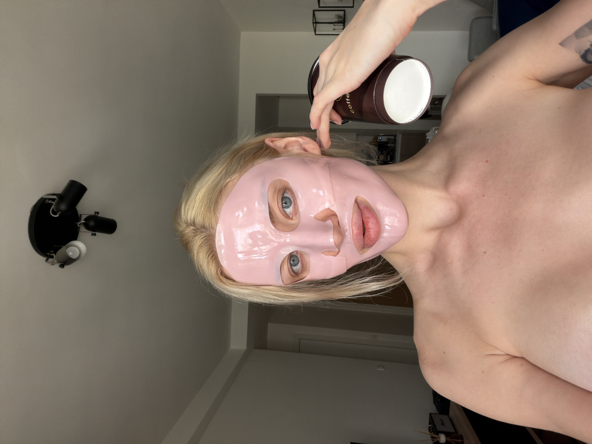 A person with a pink facial mask is visible, holding a coffee cup. The room is mostly plain, with a light fixture on the wall. The location is not discernible from the image.