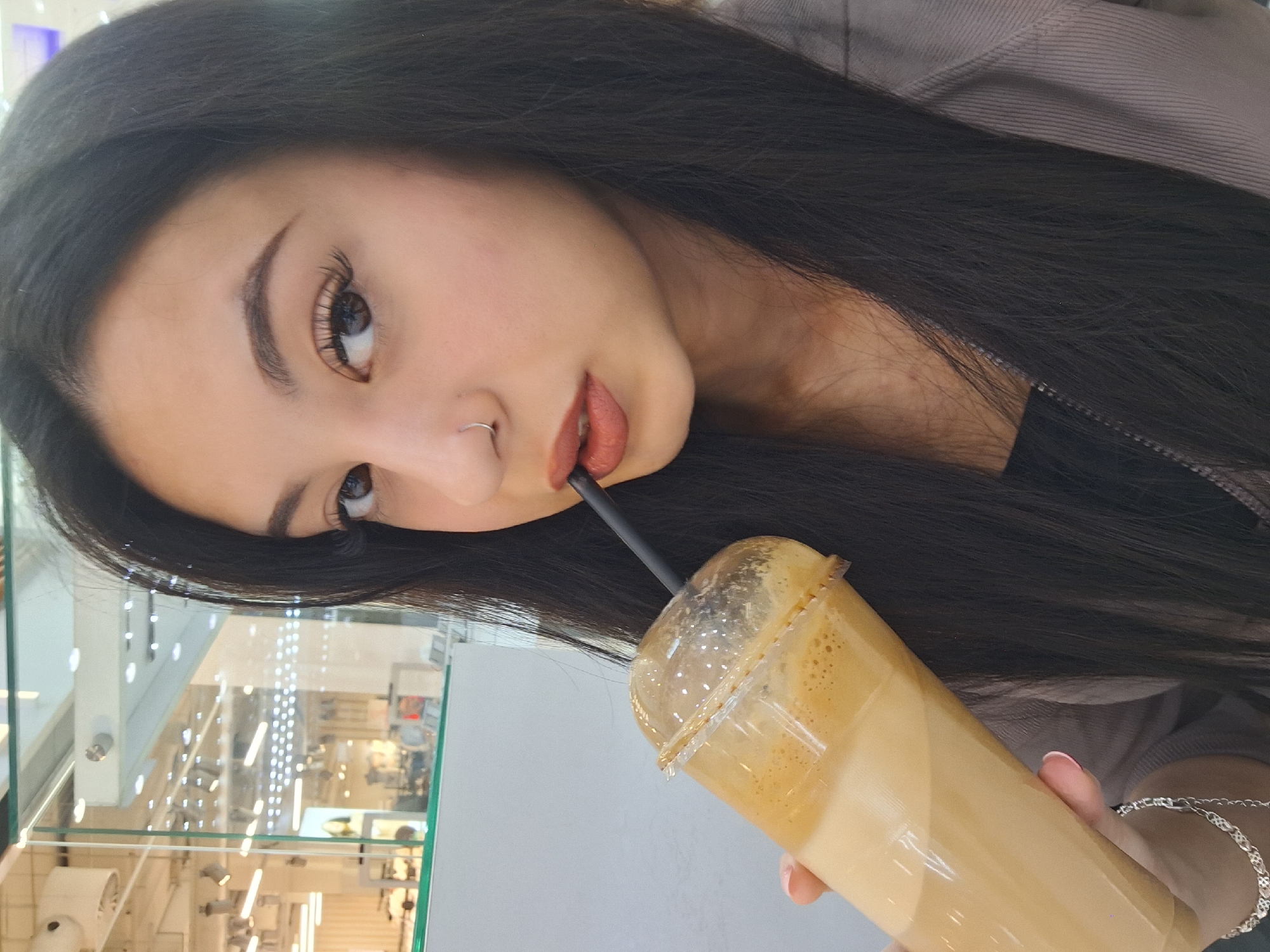 A young woman is shown holding a clear plastic cup with a beverage in a shopping mall or public space. The beverage appears to be a light tan color, possibly coffee or a similar drink, and it has some foam at the top. The woman is using a black straw to drink the beverage. She has dark hair and fair skin, with makeup on her face. She has a nose ring. The shopping center seems to be in Sofia, Bulgaria. In the background are store displays and lighting. The woman is wearing a casual top and a bracelet on her wrist.