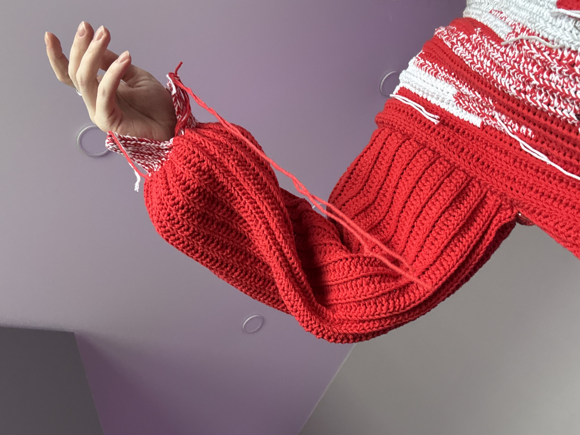 A close-up, low-angle shot shows a person's arm wearing a red and white crocheted sweater. The arm is bent at the elbow, with the hand near the upper edge of the frame. The sweater sleeve is predominantly red, with rows of visible crochet stitches. A portion of the sweater above the sleeve features alternating stripes of red and white crochet. Loose red yarn strands hang from the sleeve. The person's hand is visible, with slightly extended fingers. The background is a light purple or lavender wall, possibly a ceiling. Two circular recessed lights are visible in the background. The location is unknown and cannot be identified as Krasnoyarsk, Russia from the image.