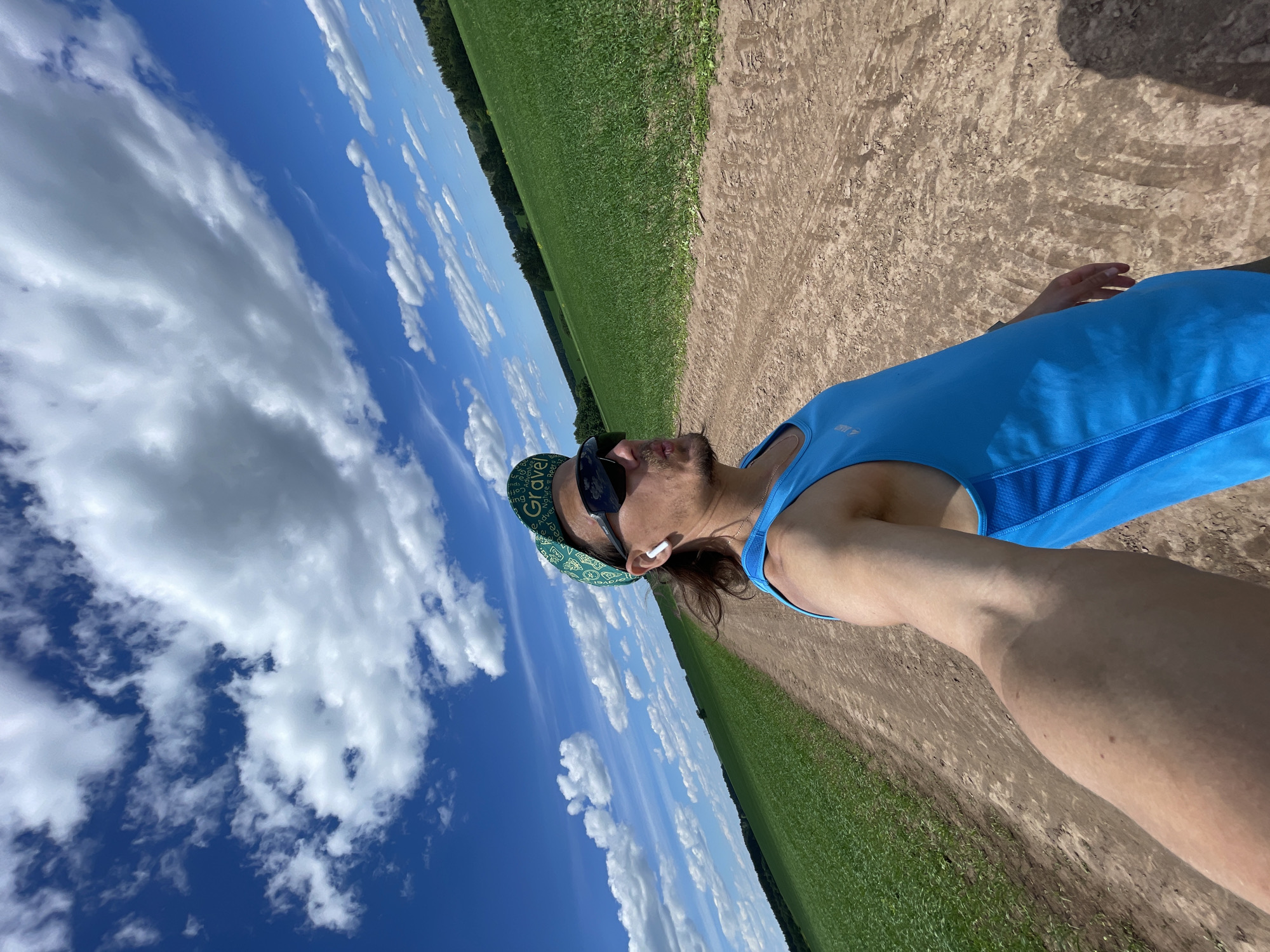 A single male individual is captured in a wide-angle selfie in a rural setting, Unknown, Unknown. He is wearing a bright blue athletic tank top with mesh panels, dark wraparound sunglasses, and a green bandana or cap with white text (partially visible: "Gravel," "Adventure," "Beer," "Nature," "Ride"). A white wireless earbud is visible in his right ear. He has long brown hair, a goatee, and his lips are pursed. His right arm, holding the camera, is visible extending into the frame, showing a tan line.
The background features a wide, unpaved dirt path with visible tire tracks, extending into the distance. Lush green agricultural fields flank both sides of the path and stretch towards a distant tree line. The sky is a vibrant blue with numerous scattered white cumulus clouds. The scene is brightly lit by direct sunlight, casting distinct shadows.