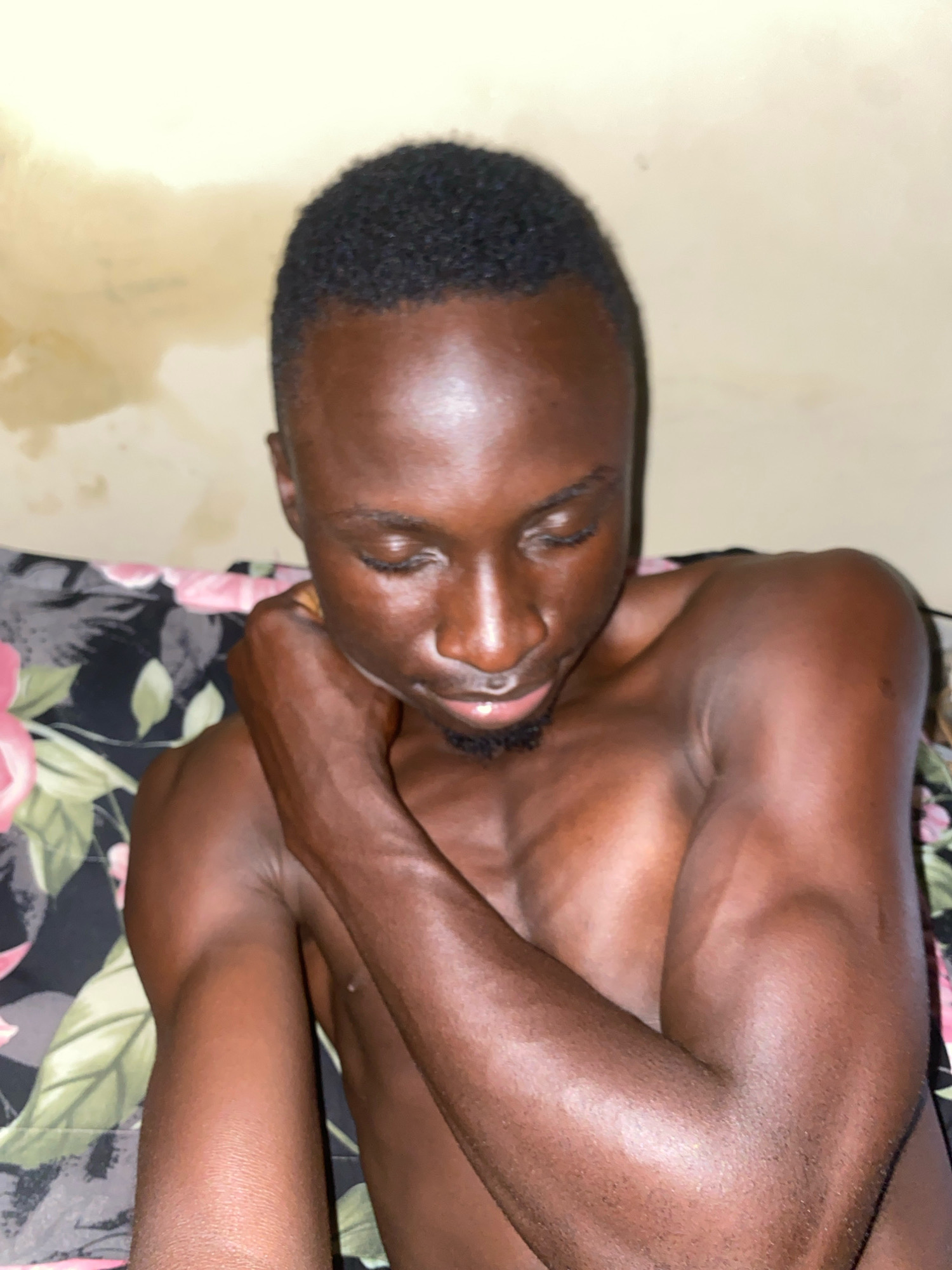 This is a close-up, waist-up, full-body portrait of a young Black male, with dark skin and short, dark hair, looking down. He is shirtless and appears to be lying down on a bed or sofa covered with a floral-patterned fabric. His left arm is bent, with his hand resting on his neck. The background is a plain, light-colored wall with some water stains. The lighting is dim, with a light source from the front. The image was taken in Barden Yana, Nigeria.