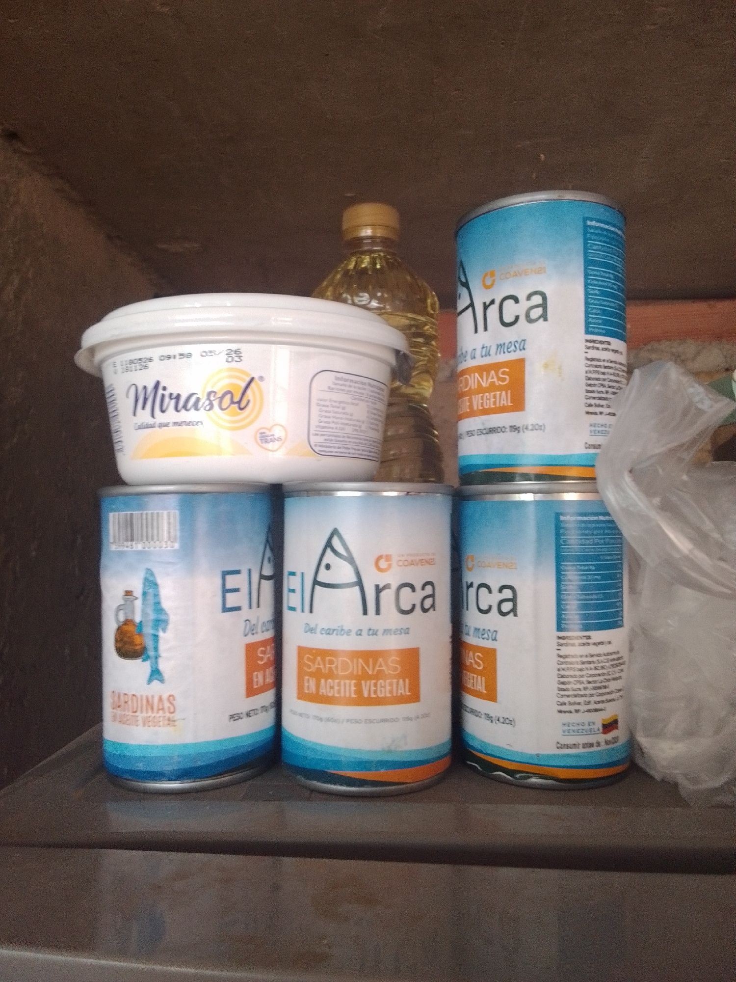 This is a medium shot photograph taken indoors, likely in a pantry or storage area, given the dark, rough, wooden background and shelves. The scene is illuminated by artificial light, with no indication of time of day or weather.
The primary subjects are several food products arranged on a shelf. On the top shelf, a white tub with a blue and yellow label is prominent, reading "Mirasol" and "Calidad que mereces". Beside it, and slightly behind, is a clear glass bottle containing a golden liquid, likely cooking oil.
Below the tub, on a lower shelf, are three cans of sardines. The two cans on the right have a blue and white label with the "Arca" logo, featuring text like "Arca... a tu mesa" and "SARDINAS EN ACEITE VEGETAL". One of these cans also displays nutritional information in a table format and indicates "HECHO EN VENEZUELA". The can on the far left also appears to be a sardine can with a similar blue and white design, featuring an illustration of a fish and bottle. This can also has text indicating "SARDINAS EN ACEITE VEGETAL".
The arrangement of the items suggests they are stored for later use, rather than being part of an active event. The overall impression is one of organized food storage, with a focus on the products themselves. The visible text confirms the origin of some products as Venezuela. There are no people or animals present in the image.