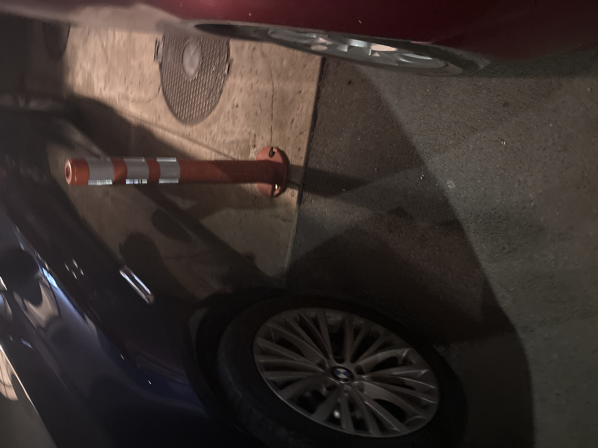 The image shows the side of a dark-colored BMW car parked on a paved surface, likely a street or parking lot, with a red car to the right. A striped orange and white traffic bollard is visible between the cars. In the background, a manhole cover with a circular pattern is set into the pavement. The scene appears to be an urban setting, possibly in Budva, Montenegro, given the presence of typical street furniture and vehicles. The lighting suggests it might be nighttime or dusk. No people are directly visible in the frame.