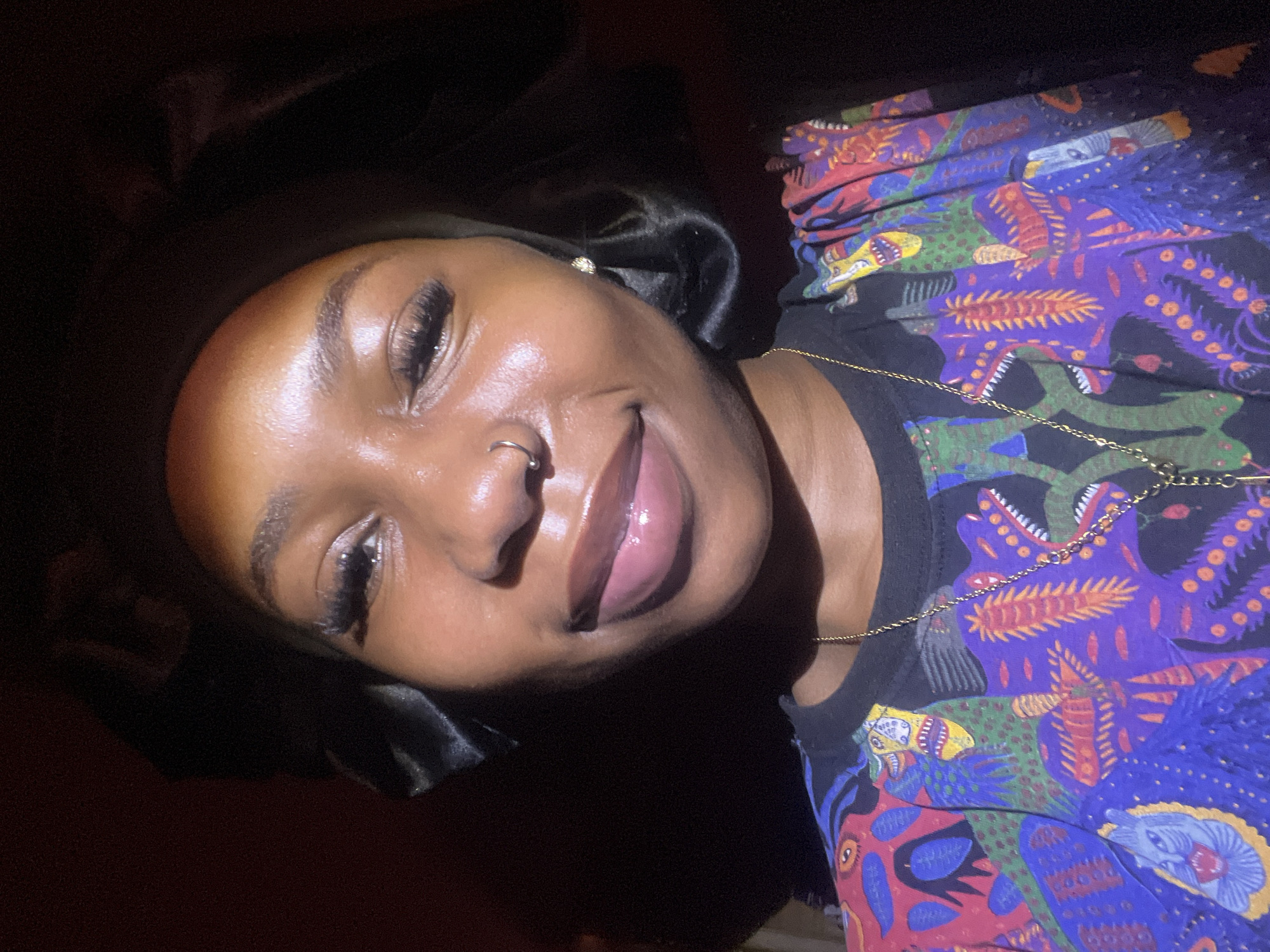 A medium shot captures a young Black woman from the chest up, facing the camera with a gentle smile. She wears a black satin bonnet, its shiny fabric pulled back to reveal her face. Her eyes are accentuated with long, dark eyelashes, and a delicate nose ring adorns her left nostril. A small, sparkling earring is visible on her right earlobe. Her lips are glossed with a soft pink hue, reflecting the light. She is wearing a vibrantly patterned t-shirt with a black collar. The shirt is adorned with a colorful, almost psychedelic design featuring stylized creatures in shades of blue, purple, red, yellow, and green. A delicate gold chain necklace rests on her chest, with a pendant visible near the center. The background is dark, with a hint of red on the left, suggesting an indoor setting. The lighting appears to be directed from above and slightly to the left, casting highlights on her face and creating shadows that add depth to the image. There is no indication of the specific location in Rafin Par, Nigeria, beyond what can be inferred from the provided context.