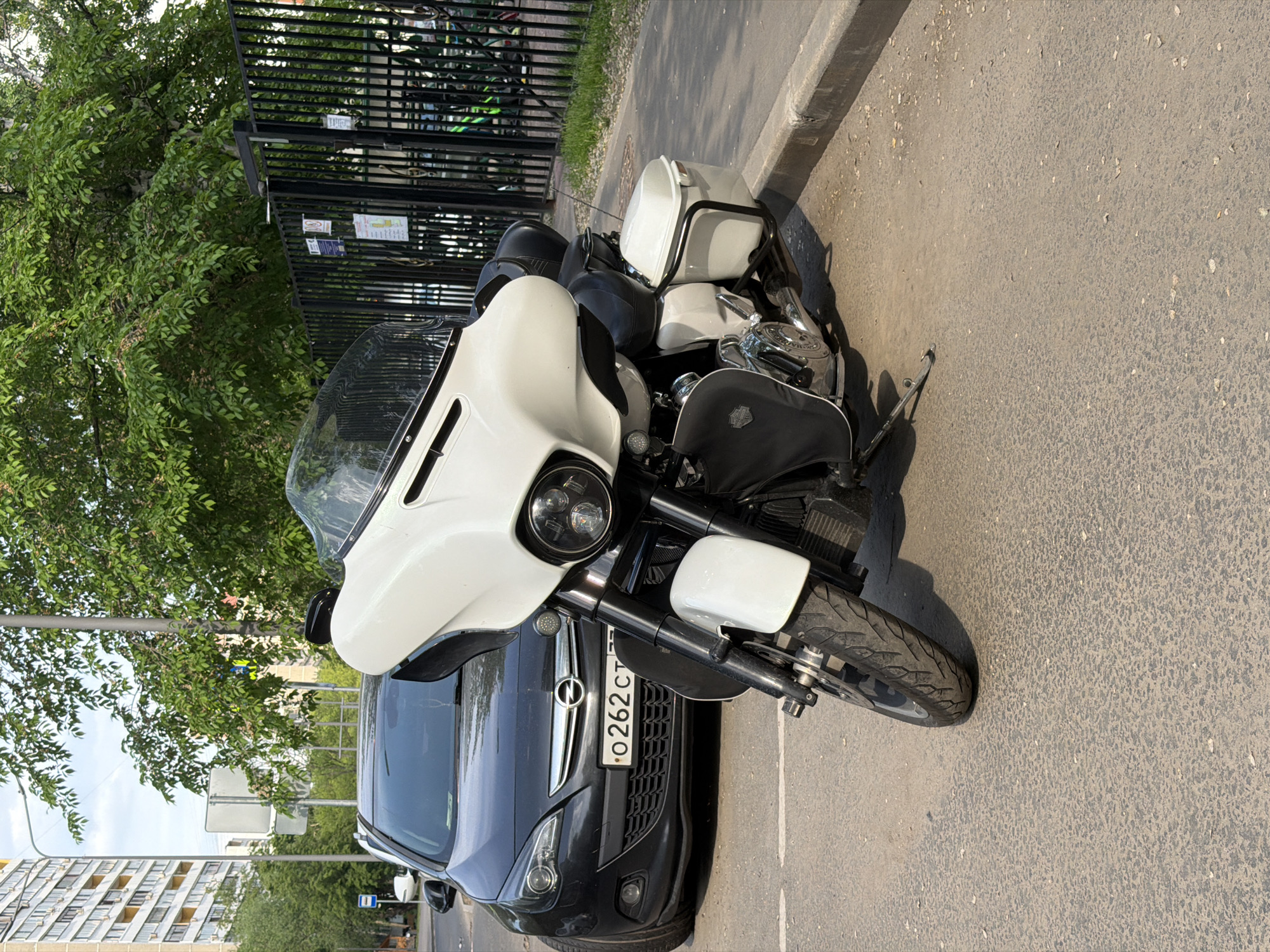 A white and black Harley-Davidson motorcycle and a dark gray Opel car are parked adjacent to each other in Moscow, Russia. The motorcycle, facing the camera, is positioned slightly forward relative to the car. The car, with a visible license plate "O 262 CT", is parked behind and to the left of the motorcycle. A tree line with a black metal fence behind it is partially visible in the upper left corner. A multi-story residential building can be seen in the lower left. The scene is outdoors, on an asphalt surface next to a curb. The lighting suggests it is daytime.