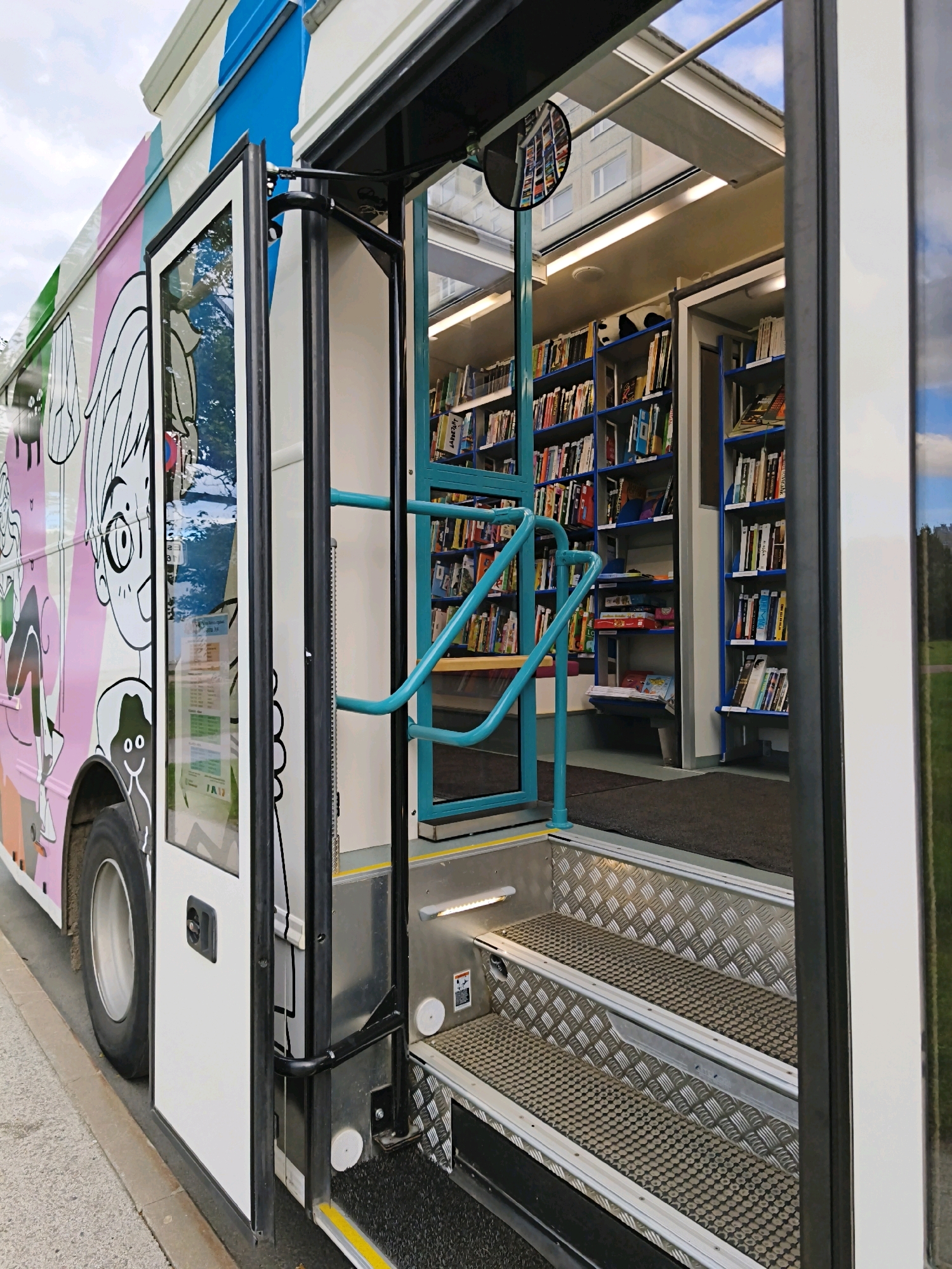 The image shows the open doorway of a mobile library bus. The exterior of the bus is decorated with colorful stripes and cartoonish drawings of people. The interior of the bus is lined with bookshelves filled with books. There are stairs leading into the bus, with silver treads and black non-slip mats. A teal-colored handrail is visible on the stairs and inside the bus. The sky is visible through the bus windows. The scene is set on a street with a sidewalk and a curb. The location is not explicitly stated in the image, but it is identified as Tallinn, Estonia in the prompt.