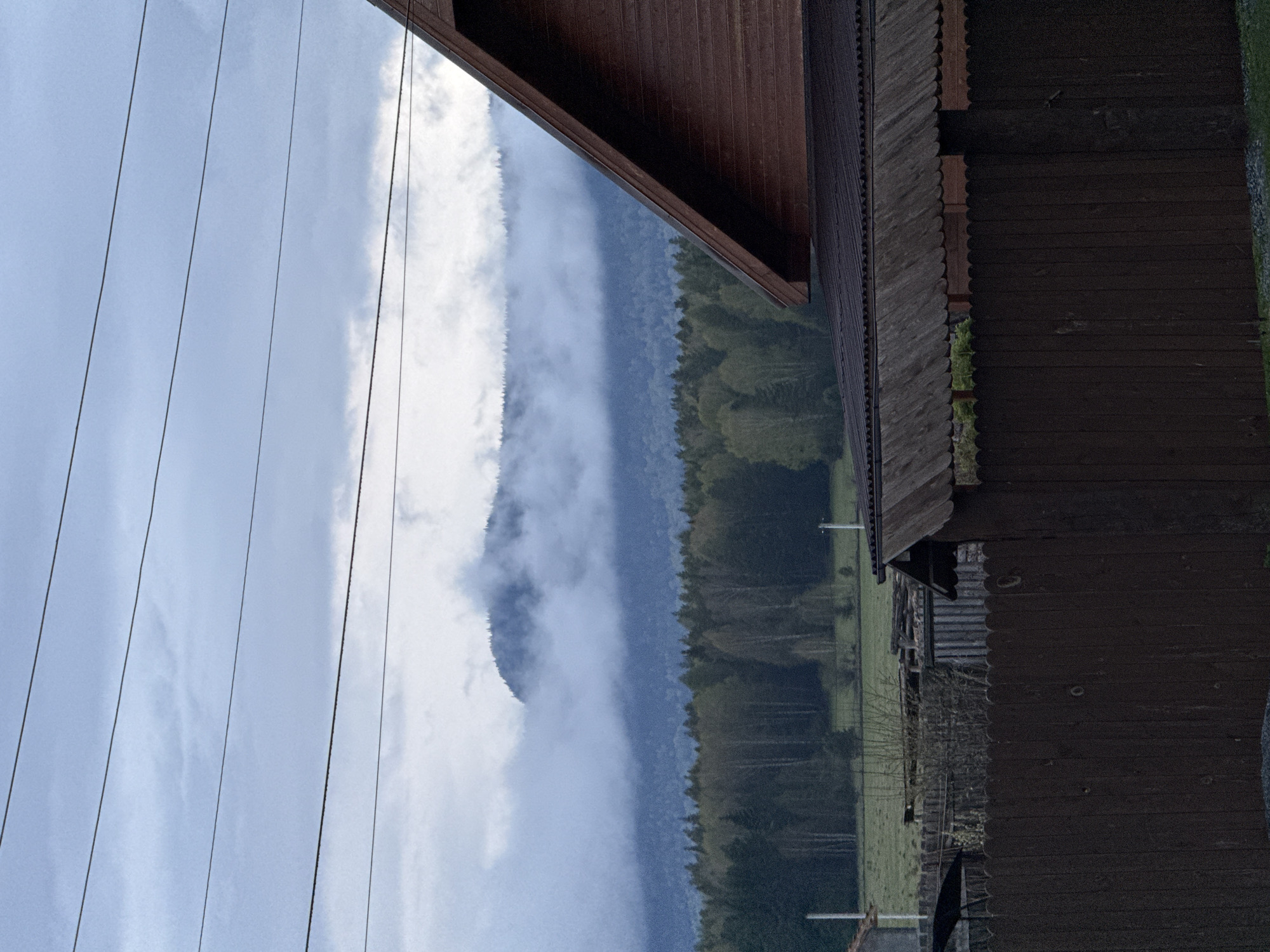 The image depicts a mountain landscape viewed from what appears to be a wooden structure. There are power lines in the upper left of the image and the roof and side of a building on the right. Mountains covered in trees are partially obscured by low-lying clouds. A green meadow is visible between the trees and the wooden structure, and power poles can be seen in the meadow.
Without additional information, it is not possible to verify the location or specific activities captured in the scene.