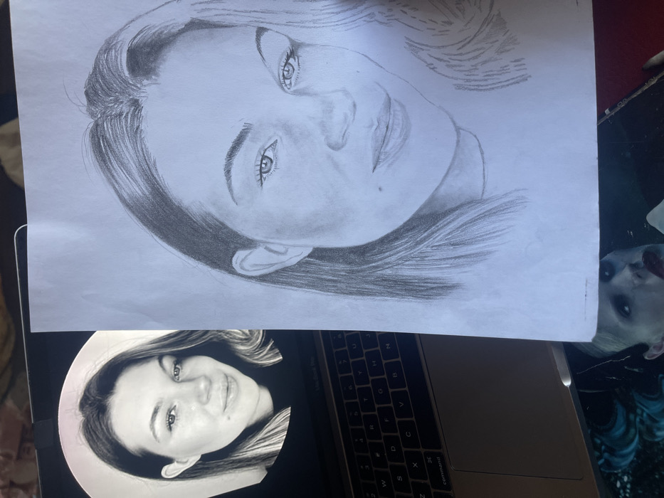 The image presents an indoor scene featuring a side-by-side comparison of a digital photograph and a graphite pencil sketch. On the left, a silver-colored laptop, identifiable as an Apple MacBook by its keyboard, displays a black and white, circular-cropped headshot of a young adult female on its screen. The woman has long, dark hair, a subtle smile, and a visible mole on her right cheek. To the right, a white sheet of paper holds a detailed pencil portrait of the same woman, rendered in grayscale, meticulously capturing her facial features, hair, and the mole, reflecting the digital reference. Partially visible beneath the laptop and paper is a dark surface, possibly a mousepad or desk mat, displaying a portion of an image depicting a face with pale skin, prominent red lips, and dark eye makeup. The environment is illuminated by ambient light. The specific location within the city of Jos, Nigeria, cannot be determined from the visual content of the image.