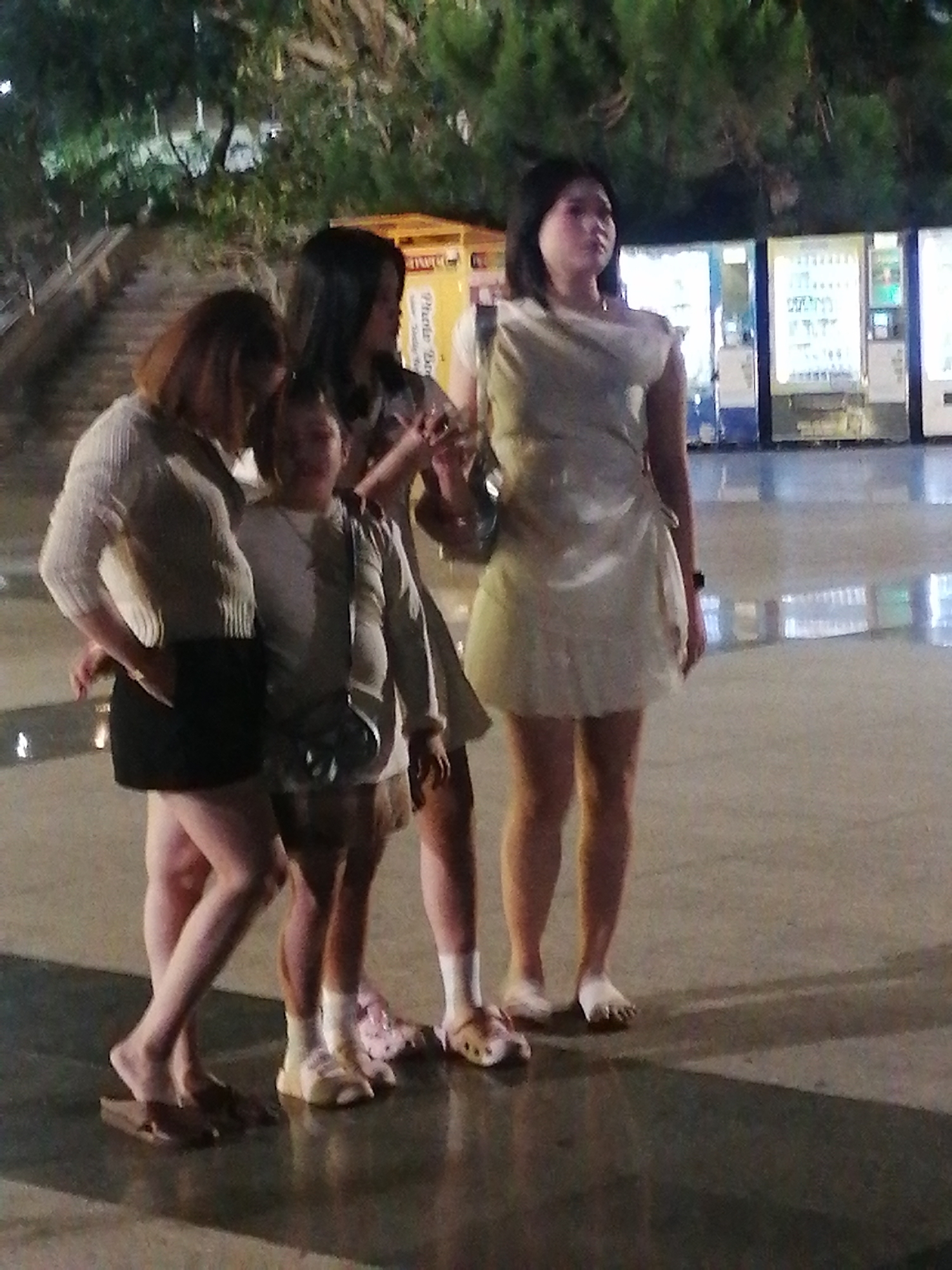 The image shows four young women standing on a wet, paved surface, possibly at night or in low light conditions. The environment appears to be an urban area with trees and buildings in the background, including what looks like a vending machine area. The women are dressed casually in various outfits. The woman on the far left wears a cream-colored, long-sleeved top and a black skirt. Next to her, a younger woman is wearing a light-colored long-sleeved shirt and a patterned skirt. The third woman from the left has dark hair and is wearing a patterned dress. The woman on the far right is wearing a light-colored, possibly satin dress. Their footwear varies, with some wearing sandals or clogs, and one appearing to be barefoot. The scene suggests a social gathering or casual outing in Da Lat, Vietnam, given the context provided.