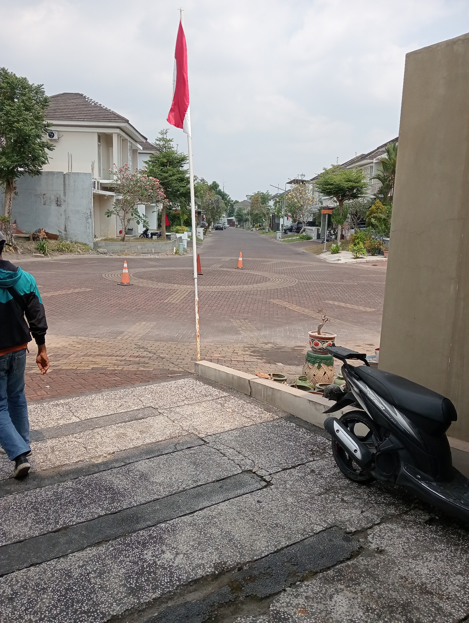 The image captures a street scene in the city of Ngaglik, Indonesia, under an overcast sky. In the foreground, a person wearing a turquoise and black jacket over blue jeans walks away from the camera, their legs and feet visible. To the right, a black scooter is parked on a textured sidewalk. The sidewalk is made of large, grey concrete slabs with a speckled pattern, interspersed with dark grey strips.
The street itself is paved with reddish-brown brick pavers arranged in a circular pattern at an intersection. Two orange traffic cones are placed on the road, indicating a potential hazard or temporary closure. A tall flagpole with the Indonesian flag, red on top and white on the bottom, stands to the left of the paved street.
On either side of the street are houses with modern architectural styles, featuring light-colored walls and tiled roofs. Trees and potted plants are visible in front of the houses, adding greenery to the residential area. In the background, more houses and trees line the street, which extends into the distance. The overall impression is one of a quiet, well-maintained suburban neighborhood.