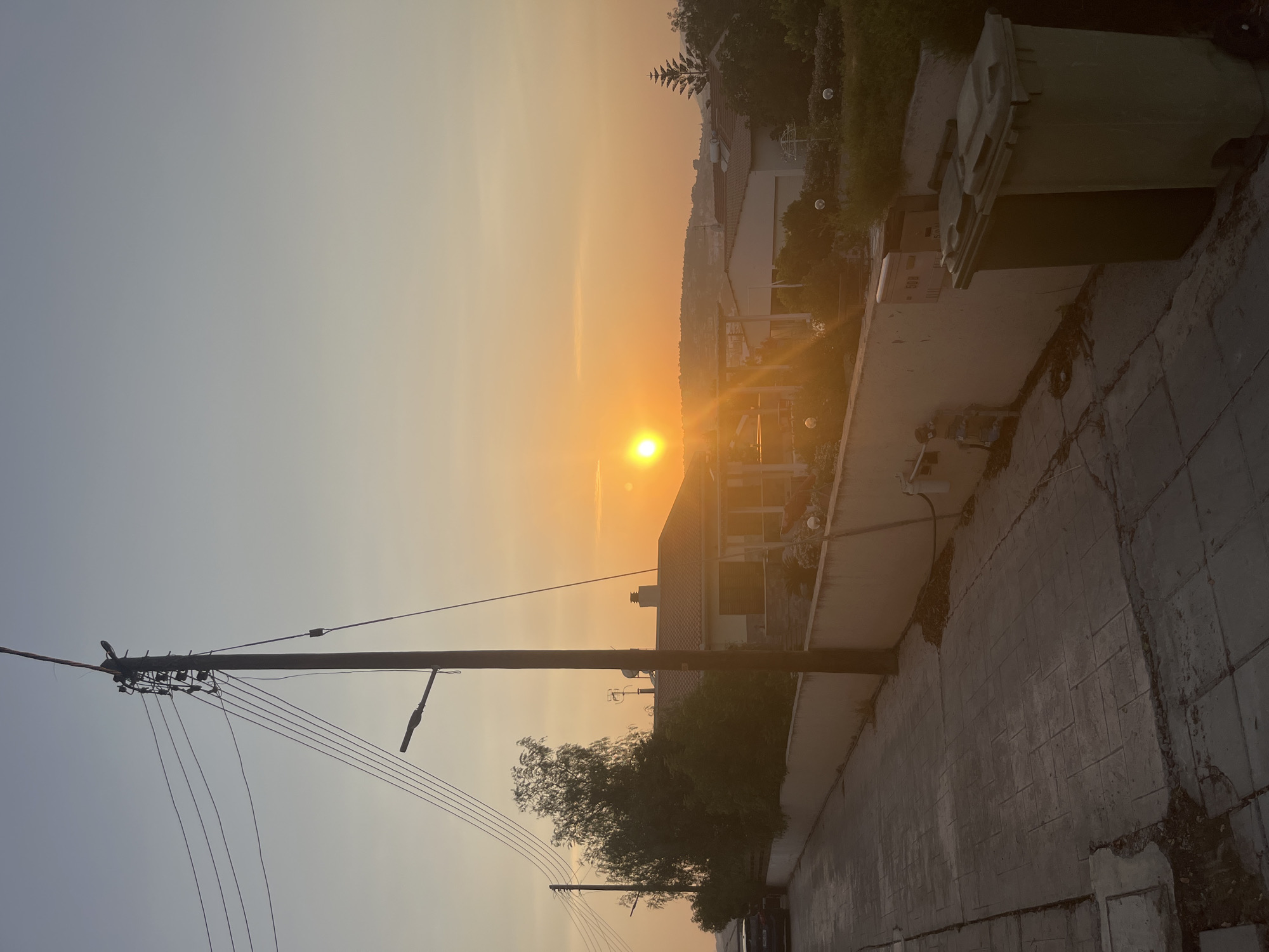 The image depicts a street scene in Parekklisia Community, Cyprus, during sunset. A utility pole with multiple wires and a street lamp stands prominently on the right side of the road. The sun, a bright orb, is low on the horizon, casting a warm orange glow across the sky and illuminating the scene.
The street is paved with stamped concrete and is bordered by a low, cream-colored wall. On the left side of the street, a dark-colored car is parked near a line of trees. Behind the wall, several houses with tiled roofs are visible, interspersed with lush green vegetation and trees. Some of the houses have balconies and outdoor seating areas.
On the right side of the paved area, adjacent to the wall, are two large, green trash bins. A mailbox with the number "508" is mounted on the wall near the trash bins. The sky transitions from a pale blue at the top to a warm orange near the horizon. The overall atmosphere is tranquil and evokes the end of the day.