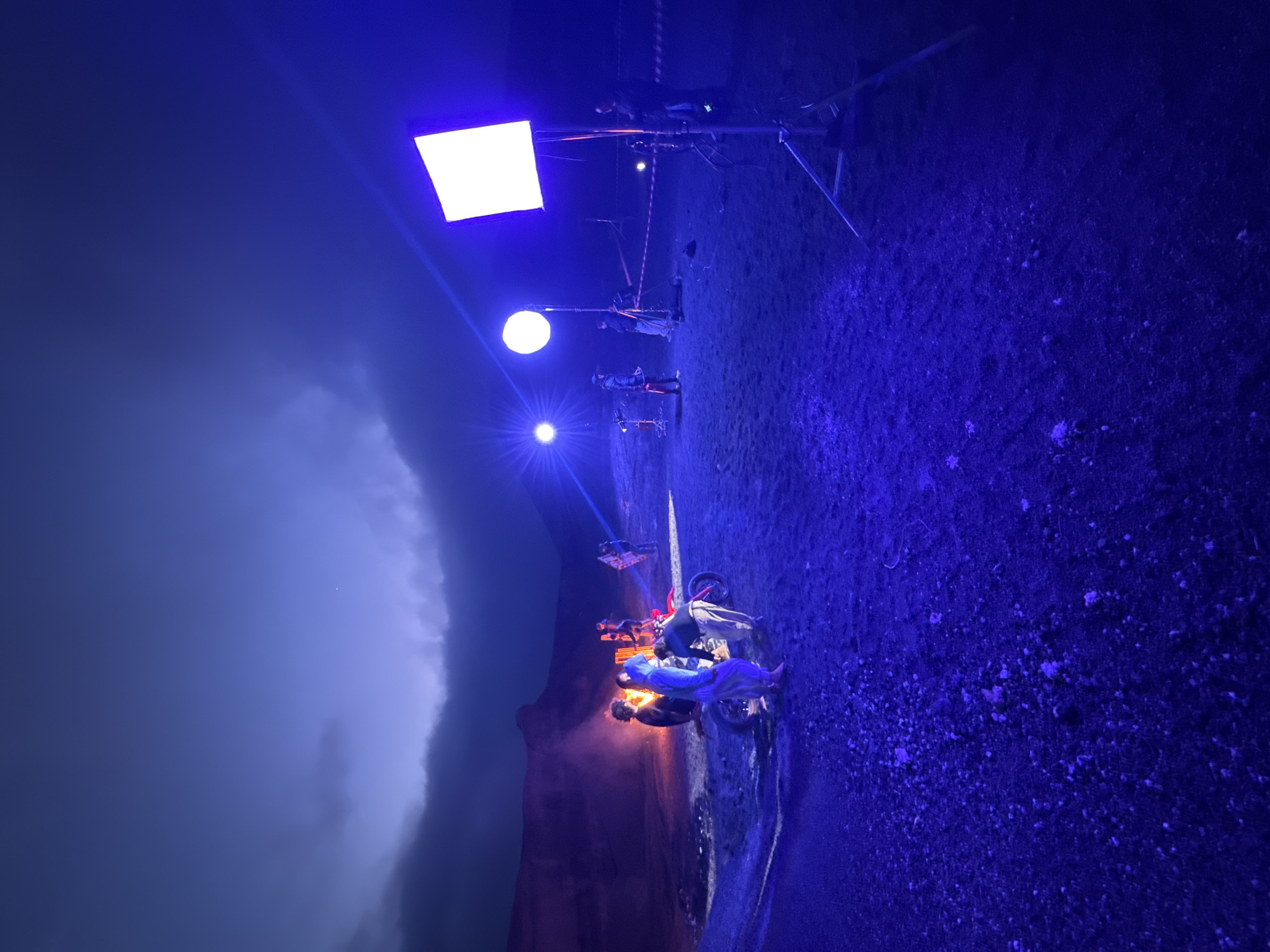 The image depicts a film set in Teguise, Spain, at night. A rocky cliff face is visible on the left, next to a shoreline. A motorcycle with two people on it is centrally located, illuminated by an orange glow, likely a fire or artificial light source. Several film crew members are positioned further back along a path, illuminated by a round light and a square panel light on a stand. The ground appears to be gravel or dirt, bathed in blue light. The sky is dark and cloudy.