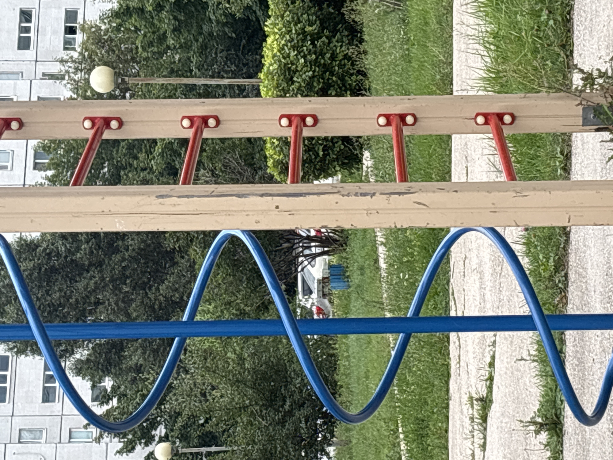 The image shows a section of an outdoor children's playground in Tolyatti, Russia. In the foreground, a blue spiral slide is partially visible on the left, next to a light brown wooden post. This post has several red horizontal bars attached to it with white caps, resembling monkey bars. In the background, there are green trees, a tall residential building with white and gray facades, and a street lamp with a spherical light. The ground appears to be a mix of grass and cracked concrete paths. There are no people or animals visible in the image. The scene is overcast, suggesting it is a cloudy day.