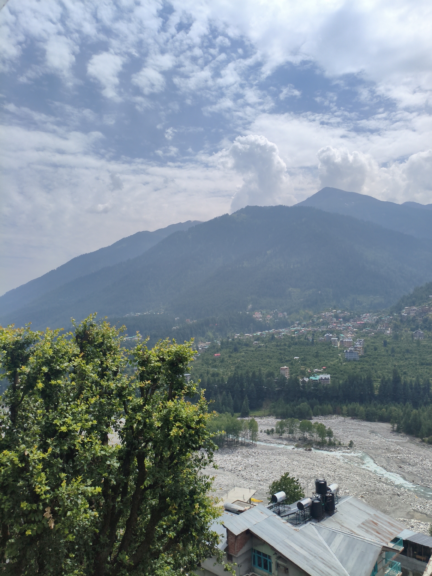 The image presents a panoramic view of the town of Vashisht, India, nestled amidst mountainous terrain under a partly cloudy sky. In the foreground, a large, leafy green tree partially obscures the view, with its branches extending towards the right side of the frame.
Below the tree, the scene unfolds to reveal a wide, rocky riverbed with a slender stream of water flowing through it. Beyond the riverbed, the town of Vashisht is visible, characterized by clusters of buildings with light-colored facades and darker roofs. These structures are spread across the slopes of a densely forested mountain that rises steeply in the background. The mountain itself is covered in a thick carpet of evergreen trees, with its peak shrouded in haze.
The sky above is a dynamic mix of blue patches and scattered white clouds, some of which are large and puffy, suggesting fair weather conditions. The overall impression is one of a serene, natural landscape integrated with a small, residential community. The image appears to be taken from an elevated vantage point, offering a broad perspective of the valley and the surrounding mountains.