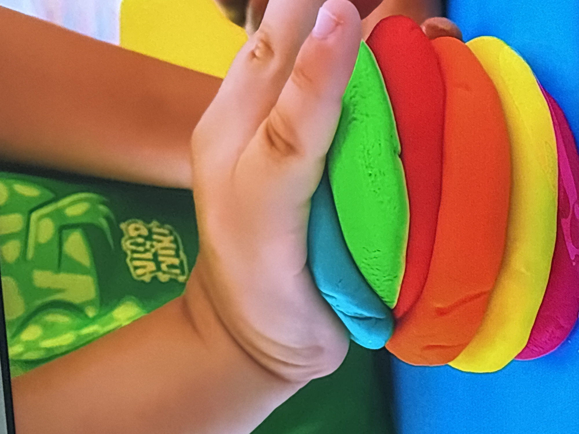 The image shows a child's hand pressing down on a stack of brightly colored play-doh layers. The play-doh is stacked in the following color order from bottom to top: pink, yellow, orange, red, green, and blue. The child is wearing a green t-shirt with a yellow graphic design and some text. The play-doh is on a blue surface. The background shows a blurred yellow object and a portion of the child's arm and torso. The image does not contain any information that would allow for the determination of its location in Dubai, United Arab Emirates.