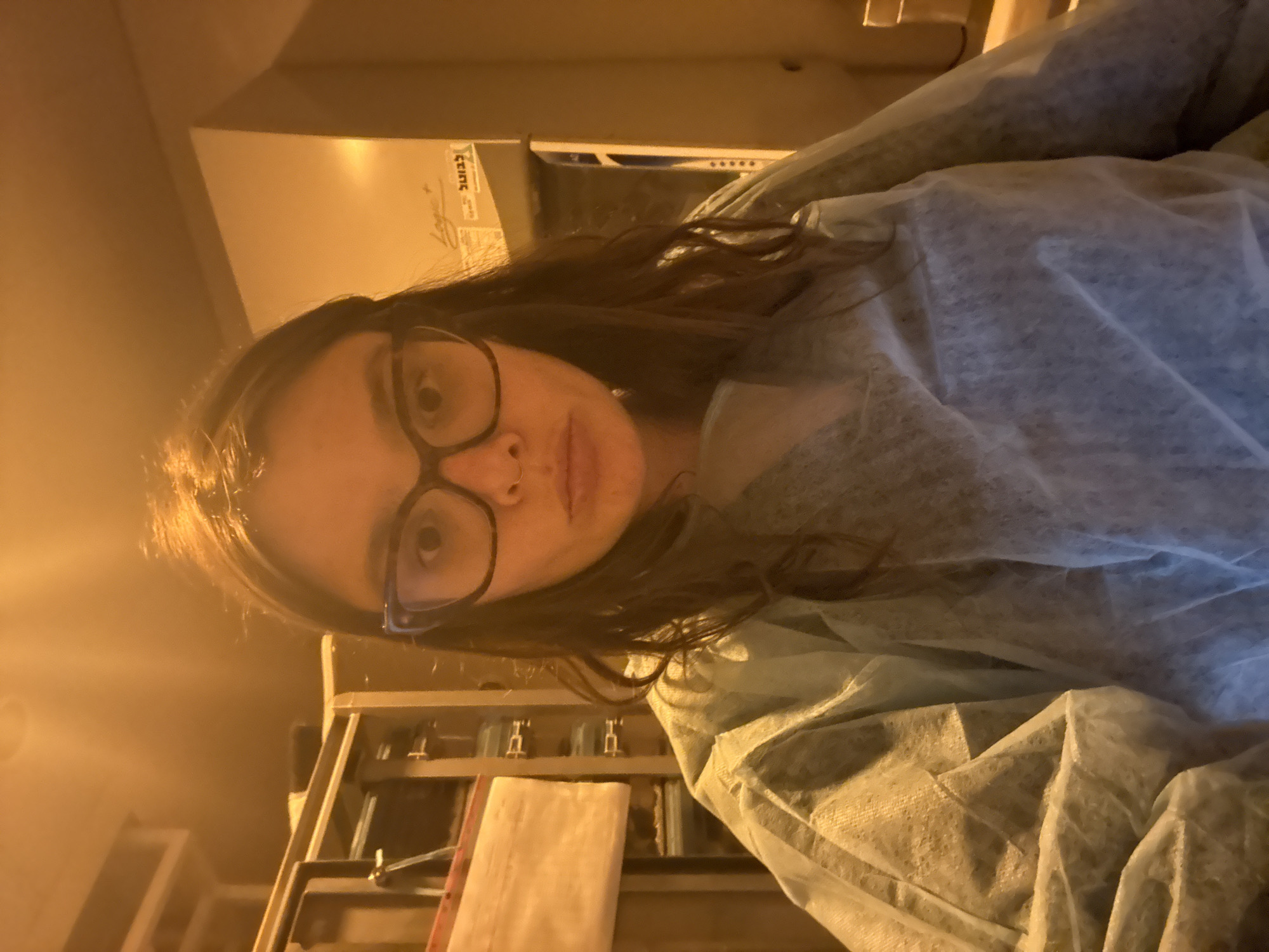 A close-up, portrait-oriented image shows a person, likely female, wearing large-framed glasses and a light blue or green disposable gown, characteristic of a laboratory, medical, or cleanroom setting. The person has dark, wavy hair and a neutral expression, looking directly at the camera. A subtle nose piercing is visible on her left nostril.
The background is warm-toned, suggesting artificial lighting, and reveals an indoor environment. On the upper right, there is a piece of equipment or a white surface with visible text. The Hebrew word "לכבוד" (L'kavod, meaning "In honor of" or "To the attention of") is discernible, along with what appears to be a signature or logo reading "Logos" below it. To the right, another Hebrew word, "בטוח" (Batuach, meaning "safe"), can be seen. Further details on this object include five small circular indicators. Below the person's head, metallic shelving or rack structures are visible, consistent with laboratory furniture. On one of these shelves, a white rectangular object is present.
The person appears to be either taking a selfie or being photographed in what seems to be a professional, controlled environment, possibly a lab or medical facility, in Tel-Aviv, Israel, as indicated by the Hebrew text. The overall atmosphere is functional and focused.