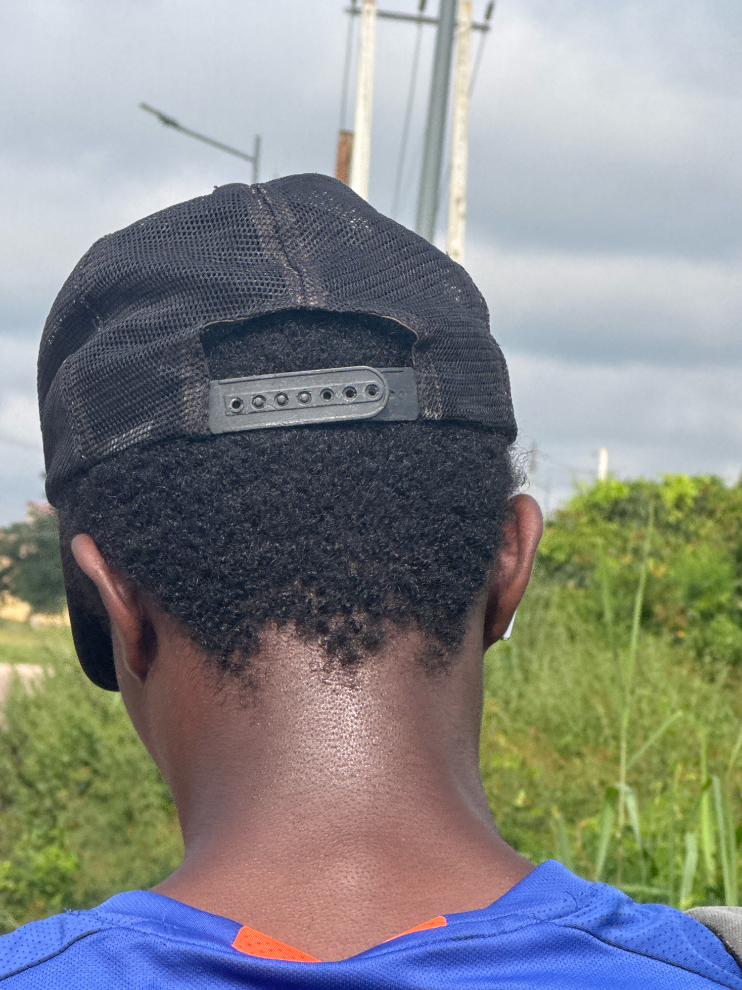 The image shows the back of a person wearing a black baseball cap and a blue shirt. The person's dark, curly hair is visible beneath the cap. The background consists of lush green foliage and a cloudy sky. The scene is likely outdoors, possibly in a rural or suburban area. The person appears to be looking away from the camera, towards the horizon. There are no other individuals or prominent objects visible in the frame. The lighting suggests it is daytime. The location is Ihiagwa, Nigeria.