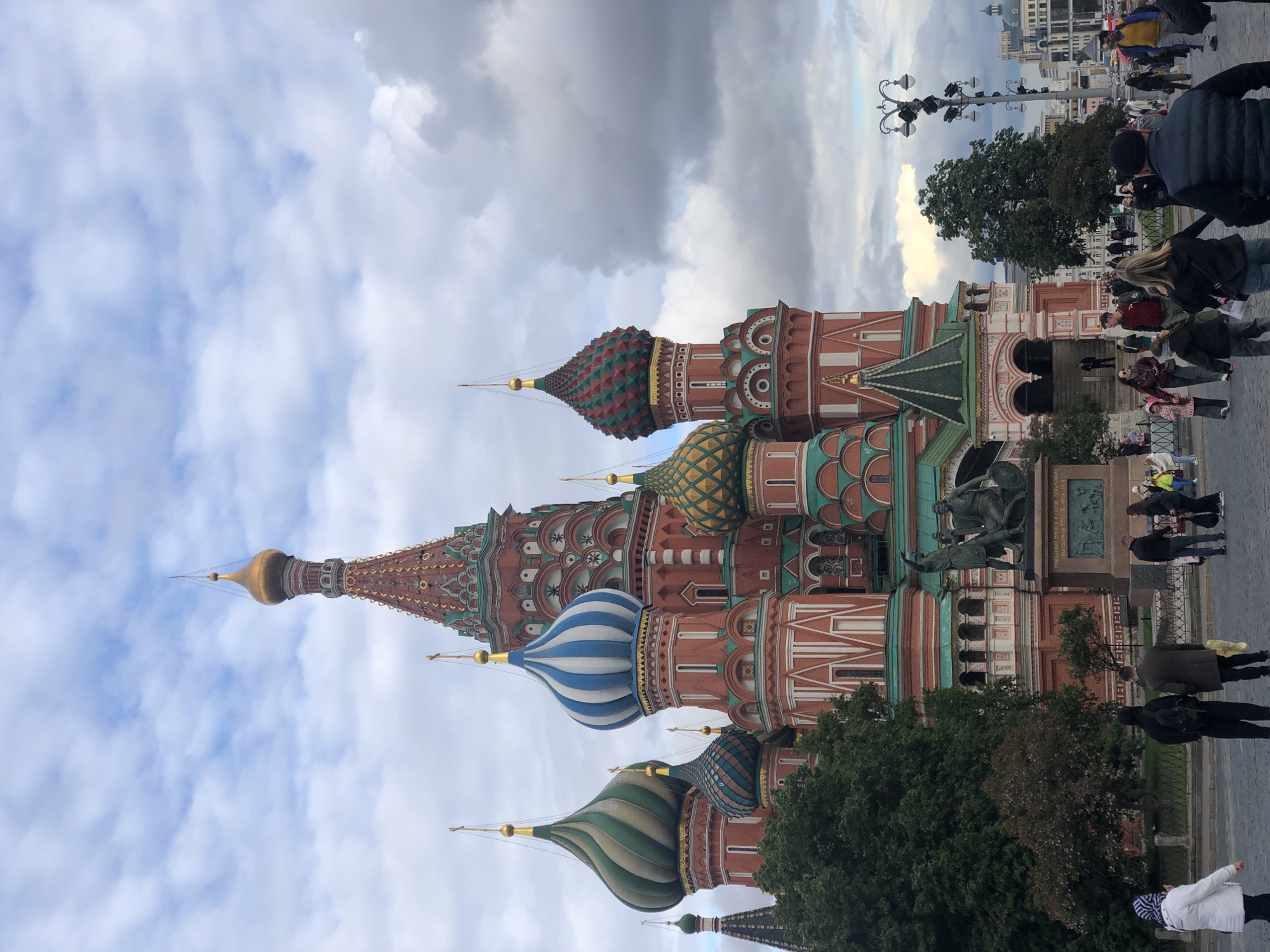 This image shows the brightly colored domes and brick facade of St. Basil's Cathedral in Moscow, Russia, against a cloudy sky. People are walking around the cathedral. There's a statue in front of the cathedral. A row of lampposts and trees is in the background, along with other buildings.