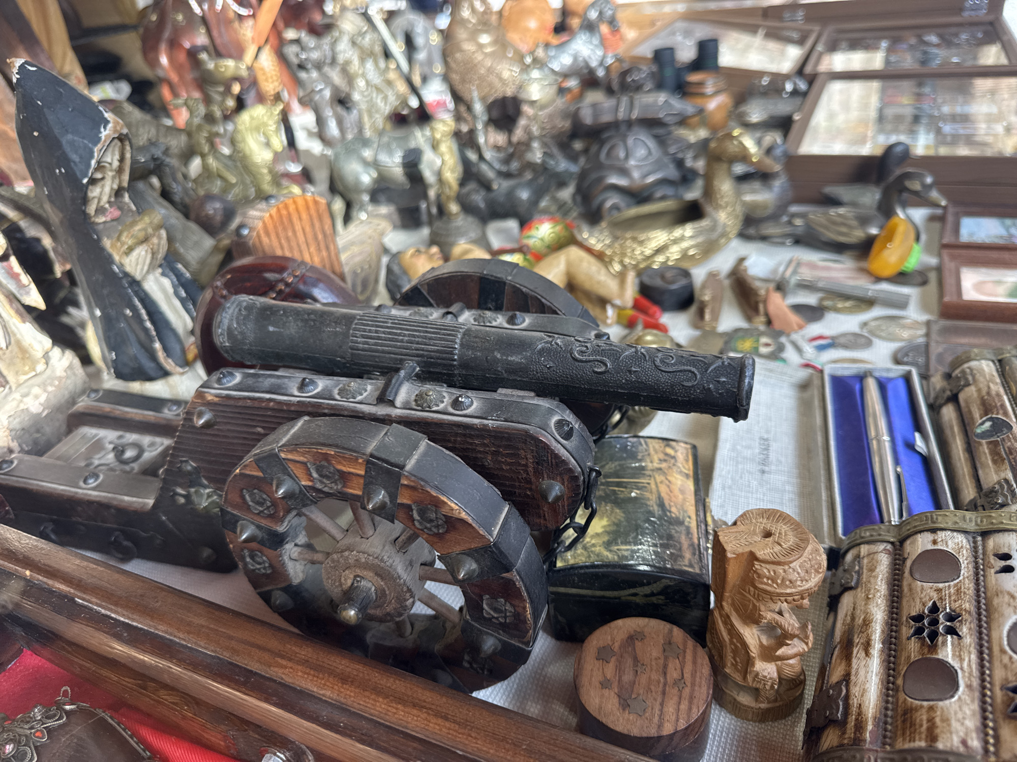 This image captures a vibrant and densely packed display of various decorative objects, likely at a market stall or an antique shop in Barcelona, Spain, as indicated by the provided context. The scene is characterized by its cluttered arrangement, presenting a treasure trove of unique items.
In the foreground, a prominent miniature cannon on a wooden carriage dominates the lower half of the frame. The cannon is dark-colored, possibly metal, with intricate designs on its barrel. Its wooden carriage features robust wheels reinforced with dark metal bands and studded embellishments, giving it an antique or handcrafted appearance.
Behind and around the cannon, a multitude of small figurines, trinkets, and boxes are crammed together. These include numerous animal figures such as brass ducks, horses, and various other stylized creatures made from different materials. Several carved wooden pieces are visible, including a small, intricately carved bust of a person and a round wooden box with star-shaped inlays. Other items comprise decorative boxes, some appearing to be made of bone or ivory with carved patterns, and one containing a silver pen set. In the mid-ground, a dark, pointed object resembling a stylized figure or ornament stands out on the left. The background is filled with more blurred objects, including display cases suggesting further collections of small items like coins, medallions, and other curiosities.
The items generally share an aesthetic of vintage, handcrafted, or ethnographic artistry, utilizing materials like wood, various metals (brass, bronze, iron-like), and possibly bone or resin. The lighting appears even, suggesting an indoor setting or a shaded outdoor stall. No legible text is visible on any of the objects. The overall impression is one of a busy marketplace filled with collectibles and souvenirs.