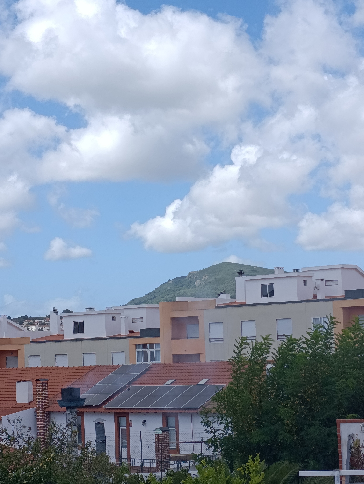 This is a photograph taken from a high vantage point, looking out over a cityscape towards a green, mountainous area under a partly cloudy sky. In the foreground, several rows of modern, multi-story buildings with flat roofs are visible. Some of these buildings have white facades, while others are painted in muted tones of beige and grey. The closest row of buildings features red-tiled roofs, one of which is equipped with solar panels. Chimneys are present on several of the roofs, some made of brick and others of a darker material. Lush green trees and foliage are abundant, particularly in the lower right and left portions of the image, obscuring some of the lower levels of the buildings. In the middle ground, a prominent green hill or mountain rises, its slopes dotted with rocky outcrops and sparse vegetation. The sky above is a bright blue, interrupted by large, fluffy white clouds. The scene appears to be a residential or urban area. The presence of solar panels suggests an emphasis on renewable energy. The architecture of the buildings, with their clean lines and predominantly flat roofs, is characteristic of contemporary construction. The mountainous backdrop provides a natural contrast to the built environment. This scene is likely from Setúbal, Portugal, a coastal city known for its proximity to the Arrábida Natural Park, which features similar mountainous terrain.