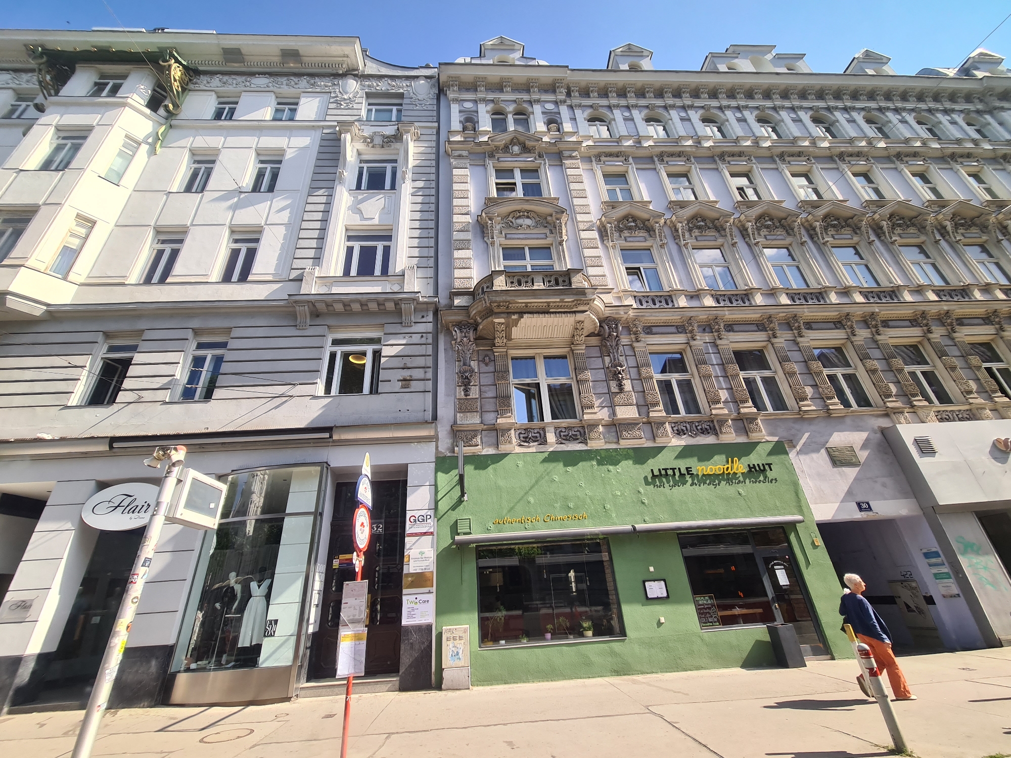 The image shows a street scene in Vienna, Austria, with two buildings facing the viewer. The building on the left is white and the building on the right is a lighter shade of gray and has ornate architectural details. In the foreground, a green building with a sign that reads "LITTLE NOODLE HUT" is visible. A person wearing an orange shirt and pants is leaning against a pole in front of the green building. The street is paved, and there are several signs and a bus stop visible. The sky is blue and clear.