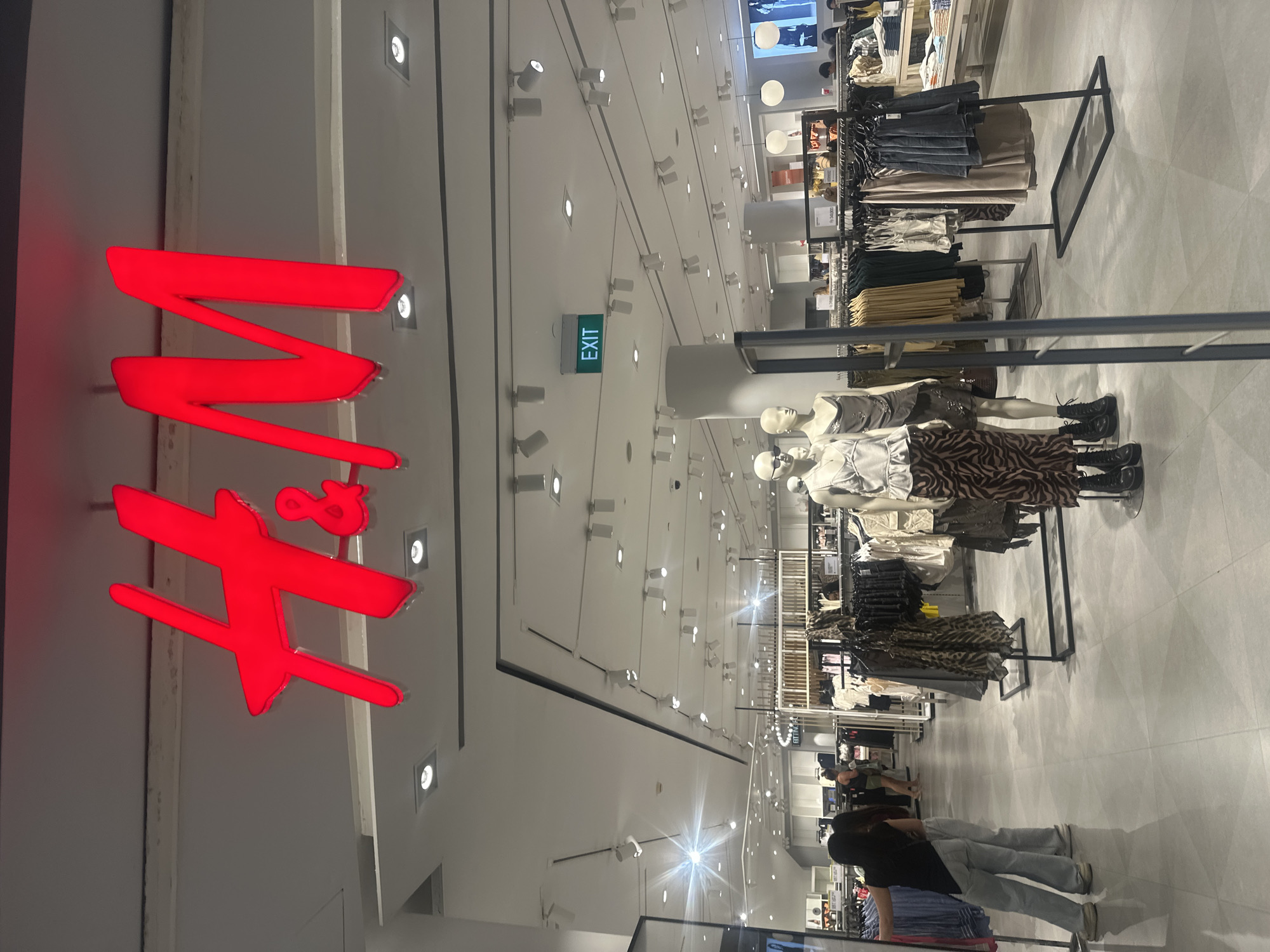 A wide shot captures the interior of an H&M store, featuring the prominent red H&M logo illuminated above the entrance. The store is brightly lit with numerous ceiling lights and displays clothing racks filled with apparel. Two mannequins are positioned in the foreground, dressed in stylish outfits. Several shoppers are visible browsing the merchandise and walking through the aisles. A green "EXIT" sign is mounted on the ceiling. The floor is tiled, reflecting the overhead lighting. The store appears to be located in Kuta, Indonesia, based on contextual information.