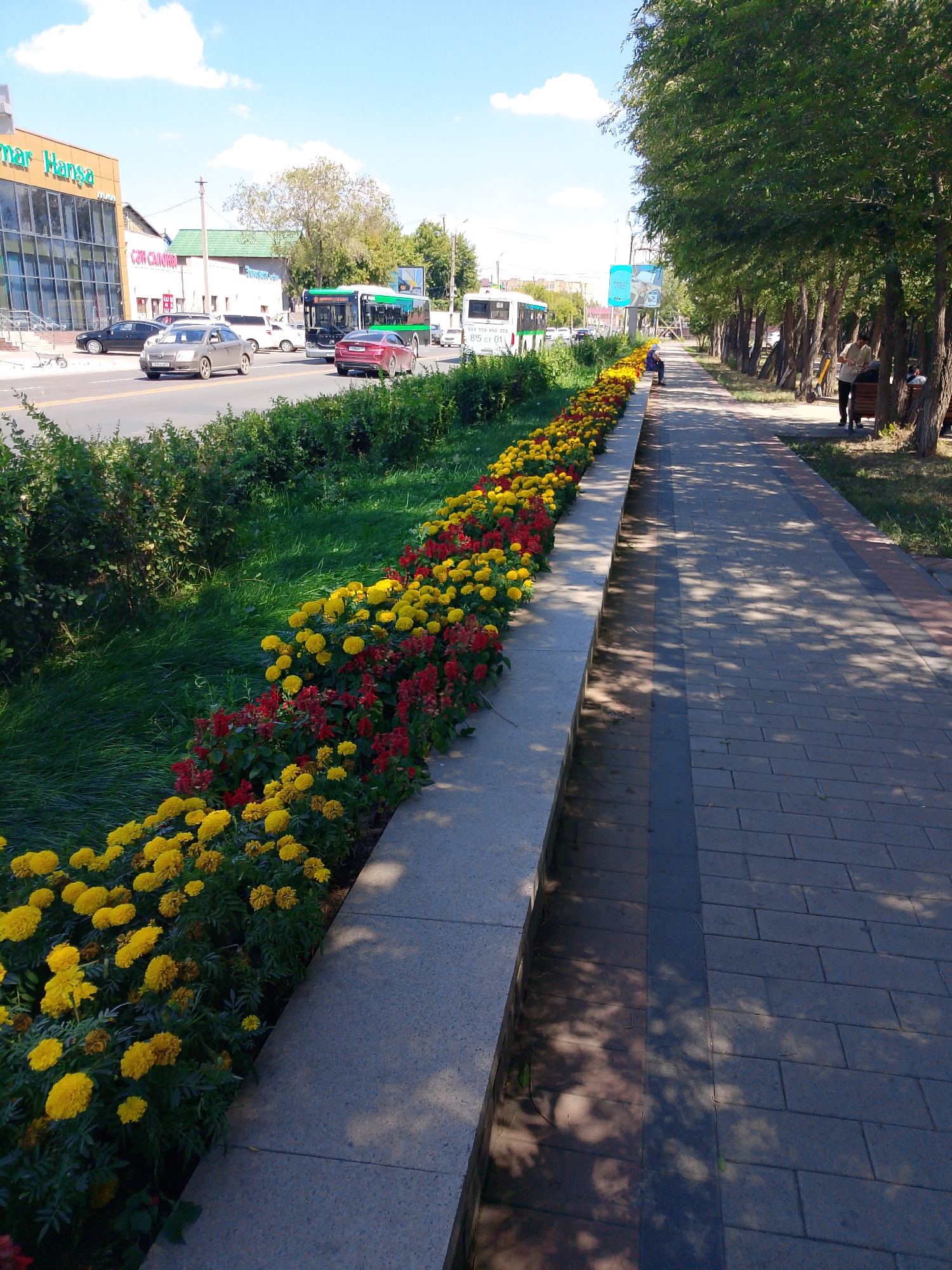 The image shows a street scene in Astana, Kazakhstan, likely along a parkway or wide boulevard. On the left, a modern building with a glass facade and an orange-colored awning is visible, identified by signage that reads "mar Hansa" in English. Adjacent to this building is a smaller structure with a green roof. The street itself is busy with traffic, including several cars, two white and green buses with visible route numbers, and a red sedan. A prominent feature of the scene is a long, raised flower bed running parallel to the sidewalk, filled with vibrant yellow and red flowers. The sidewalk, made of interlocking gray bricks, is lined with mature trees that cast dappled shadows on the path. Several people are present; one person sits on a bench along the sidewalk, another stands near a bicycle rack, and a third individual is partially visible near the flower bed. The sky is blue with scattered white clouds, indicating a clear day.