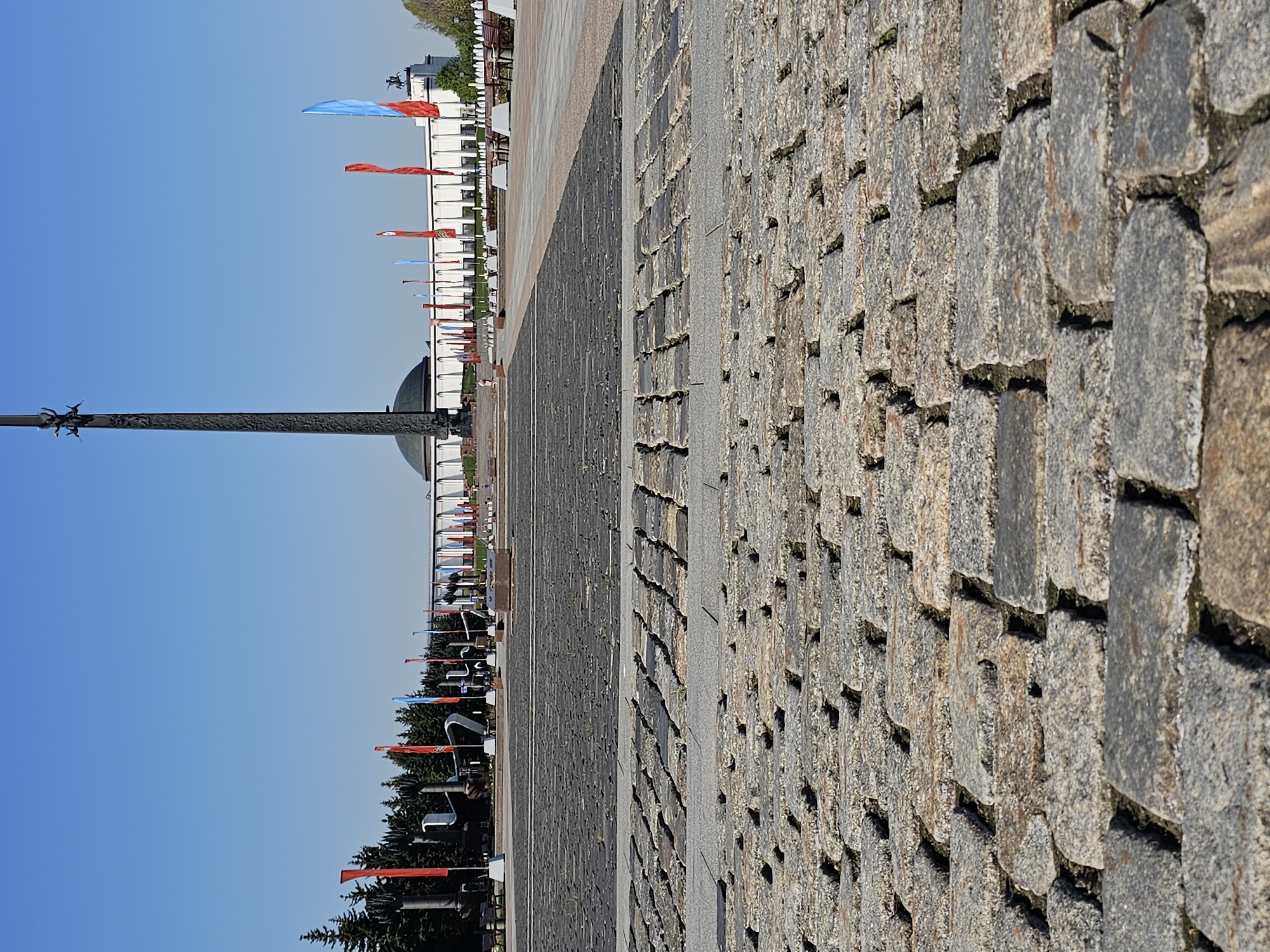 The image depicts a stone-paved walkway, likely a part of a public space in Moscow, Russia. On the left side, a tall monument is visible, topped with a statue. A domed building is in the background. Red and blue flags are seen along a row of white structures that could be part of a memorial or building complex. Trees line the background. The sky is clear and blue. The image appears to be taken during the day with good lighting.