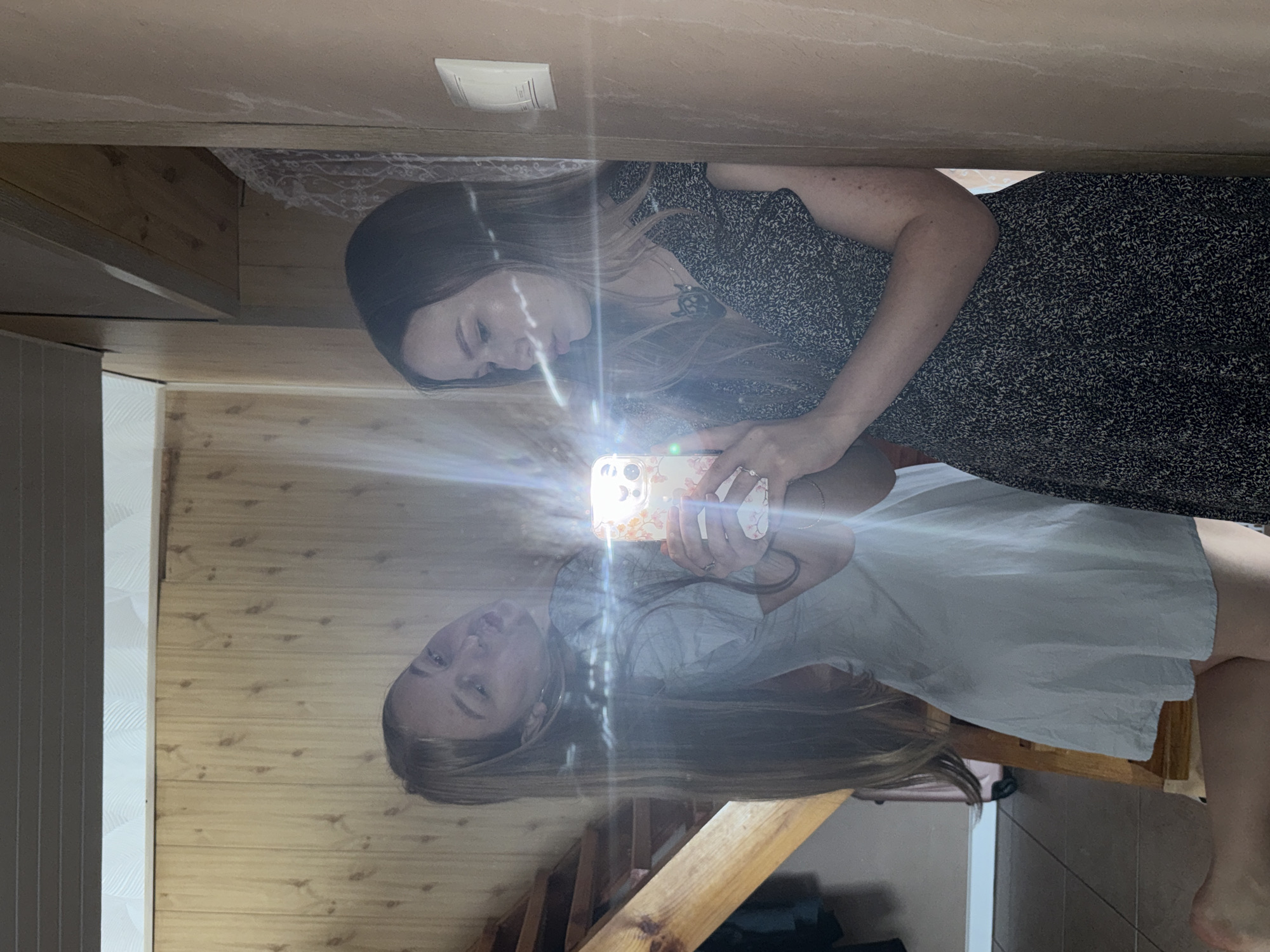 Two young women pose for a mirror selfie in a room with wood-paneled walls and a staircase. The woman on the left, reflected in the mirror, wears a light blue dress and has long blonde hair. She leans into the frame with her head tilted. The woman on the right, standing in front of the mirror, wears a dark patterned dress. She holds a smartphone with a floral case, capturing the reflection of her friend and herself. The room appears to be part of a house or cabin, possibly in Zhigalki, Russia, given the rustic wood elements and simple interior.