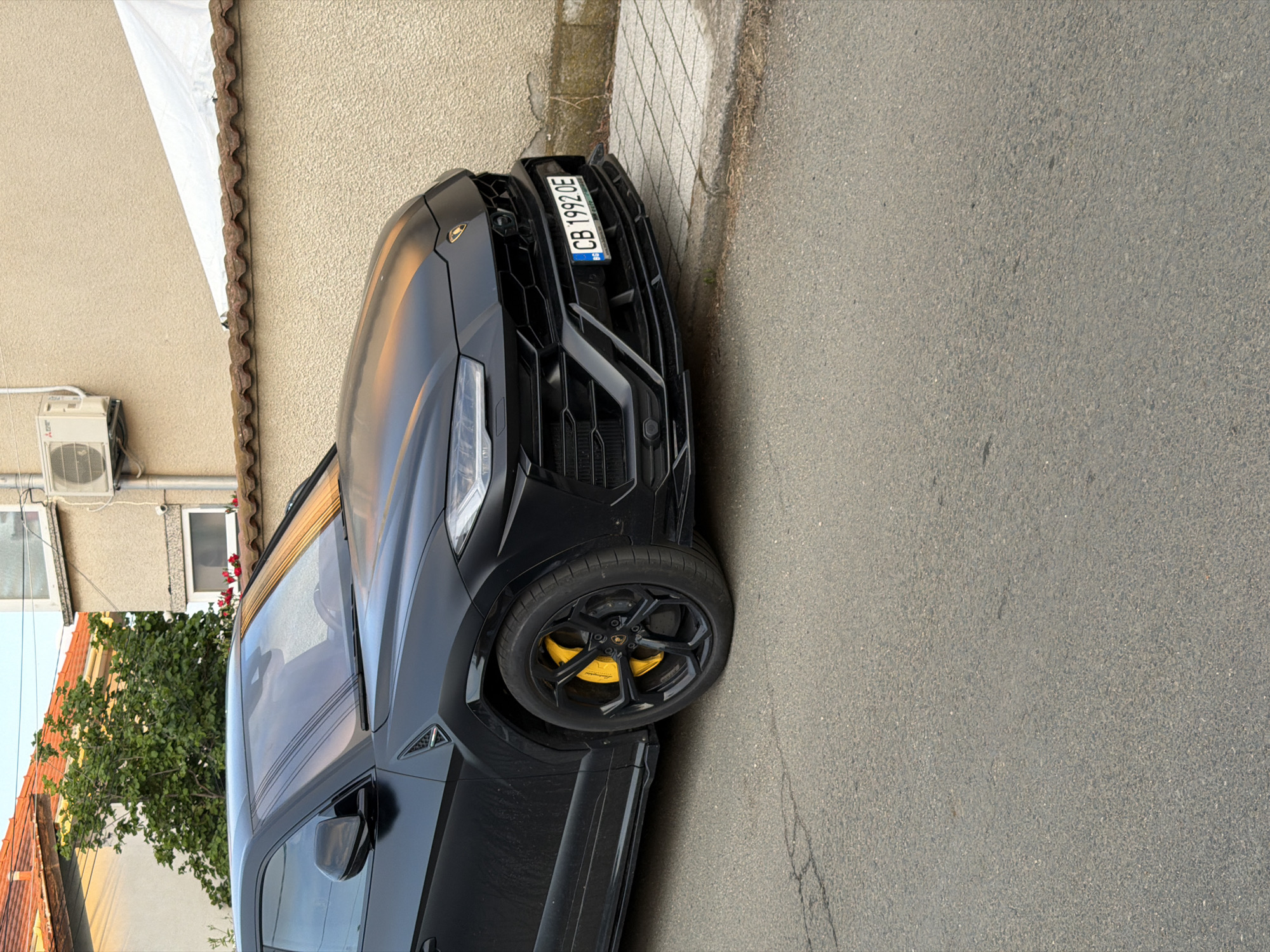 The image shows a black Lamborghini Urus parked on a paved street next to a sidewalk. The car's license plate reads "CB 1992 OE". The front of the car is visible, including the yellow brake calipers. A building with a beige wall and a white overhang is in the background, along with some greenery. Based on the license plate and general architectural style, the location is likely in Burgas, Bulgaria. There are no people visible in the image. The weather appears to be overcast.