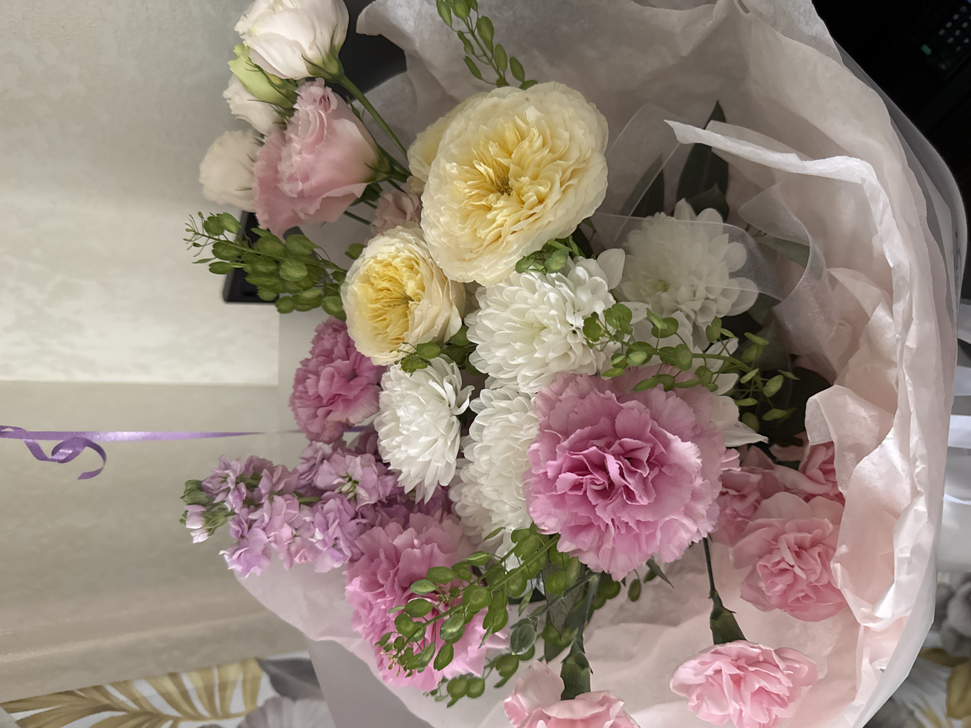 A bouquet of flowers wrapped in tissue paper sits indoors. The flowers are a mix of colors including white, yellow, pink, and lavender. There are roses, carnations, chrysanthemums, and stock flowers visible. A light purple ribbon is draped across the top left corner. The location is unknown, and there is no evidence it is in Sochi, Russia.