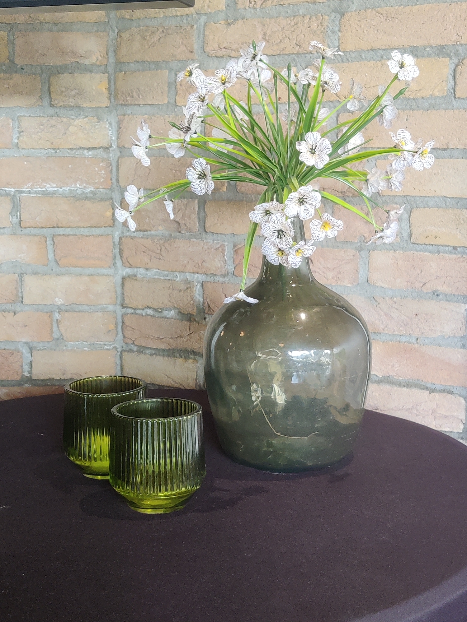 The image displays a still life composition with a brick wall in the background. In the foreground, on a dark, textured surface, are two green ribbed glasses and a large, dark green glass bottle containing a bouquet of white artificial flowers. The glasses are identical and positioned side-by-side. The bottle is bulbous at the bottom and tapers towards the neck, with the flowers and their green stems arranged within it. The brick wall behind the objects is made of light-colored bricks with grey mortar. There are no people or animals visible in the image. The scene is indoors, likely in a home or a decorative display area in Winterswijk, Netherlands, given the provided context.