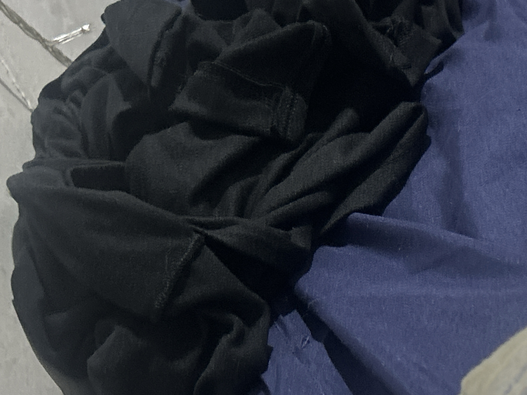 The image shows a close-up, high-angle shot of black and blue fabric. The black fabric appears to be a piece of clothing, possibly a t-shirt or sweater, folded and draped over a blue surface. The fabric is textured and creased, with visible seams in some areas. The blue surface beneath the black fabric is smooth and appears to be another piece of fabric, perhaps a blanket or bedsheet. The lighting is dim, casting shadows and highlighting the textures of the fabric. There is no discernible background or context that would indicate a specific location in Santa Ana, Venezuela.