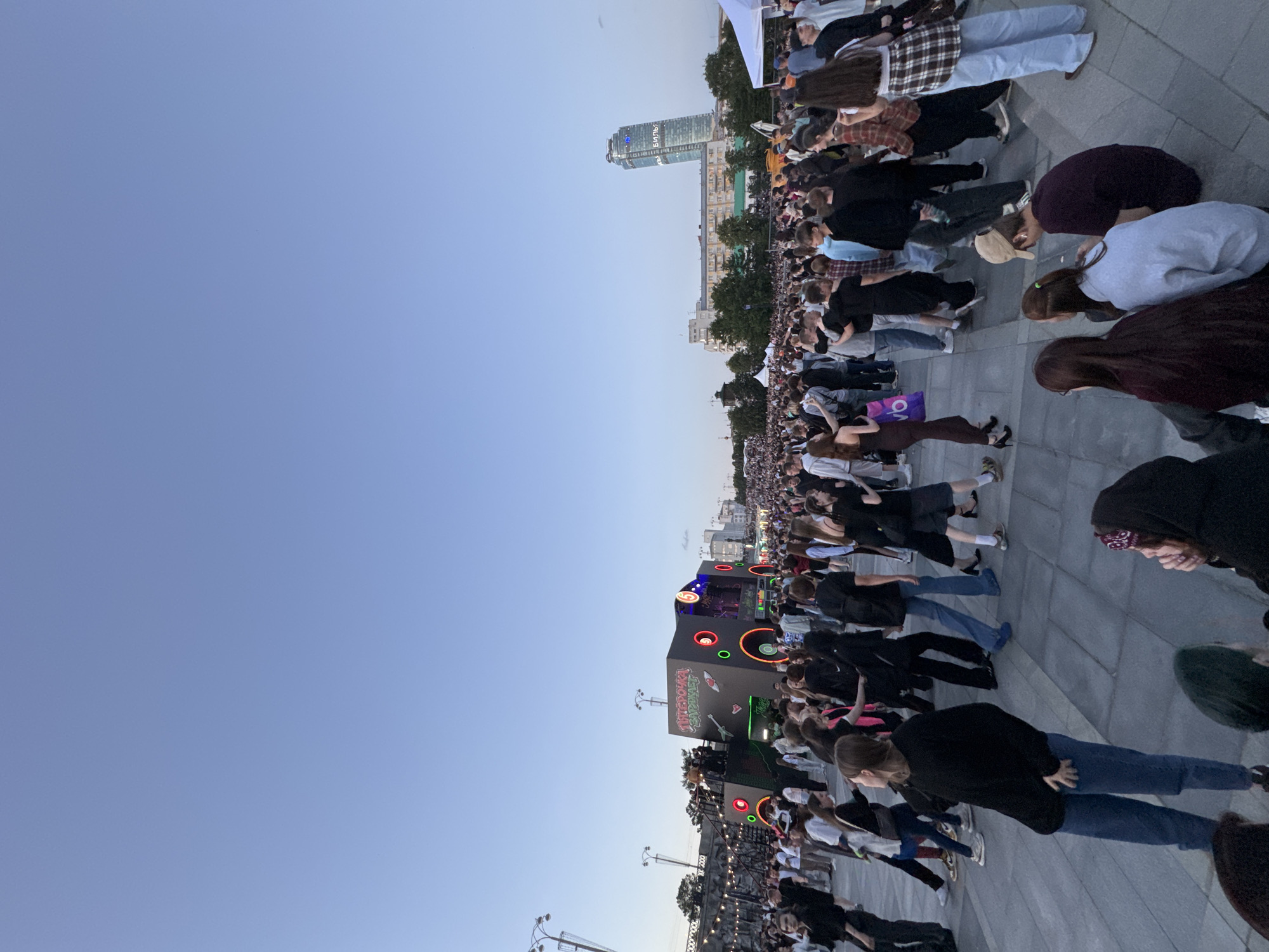 A large crowd of people are gathered outdoors in a city square during twilight. In the background, a modern skyscraper with illuminated text and other urban buildings are visible. The main focus of the scene is a stage set up with colorful lights and what appears to be large speaker structures. People are standing and walking around, some are interacting with each other, and others appear to be watching the stage. The overall atmosphere suggests an outdoor event, possibly a concert or festival. The image is taken from a slightly elevated perspective, looking down at the crowd. Based on the skyscraper with illuminated text and the surrounding architecture, this scene is likely located in Yekaterinburg, Russia.