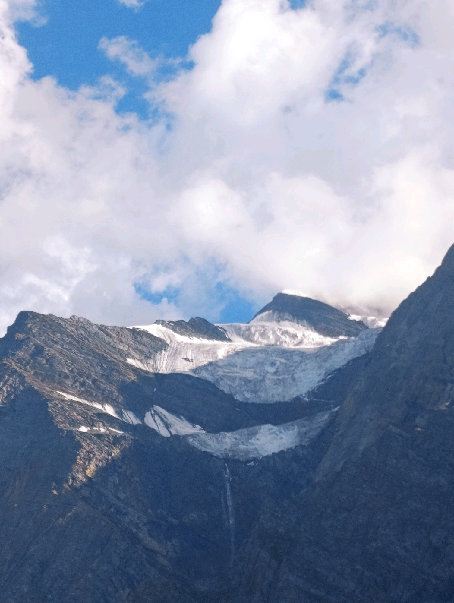 The image depicts a majestic mountain landscape dominated by a vast glacier. The glacier, a massive expanse of white ice, is situated on the upper slopes of rugged, dark grey mountains. The mountain peaks are jagged and imposing, with some exposed rock faces and patches of snow scattered across them.
The sky above is a vibrant blue, dotted with fluffy white clouds. The clouds are illuminated by sunlight, creating a sense of depth and contrast. There is a thin veil of mist or fog hovering near one of the mountain summits, partially obscuring the rock face and adding an ethereal quality to the scene.
A thin waterfall is visible cascading down the mountainside, originating from the melting glacier. The water flows downwards through the rocky terrain, eventually disappearing from view.
The lighting suggests it is daytime, with sunlight casting shadows on the mountains, highlighting the textures and contours of the rock formations. The overall impression is one of natural grandeur and the raw power of alpine environments.
The image does not contain any people or man-made objects, except for a very small, dark object that appears to be a helicopter flying near the glacier. The location in the city of Gondhla, India, is not visually discernible within the provided image.