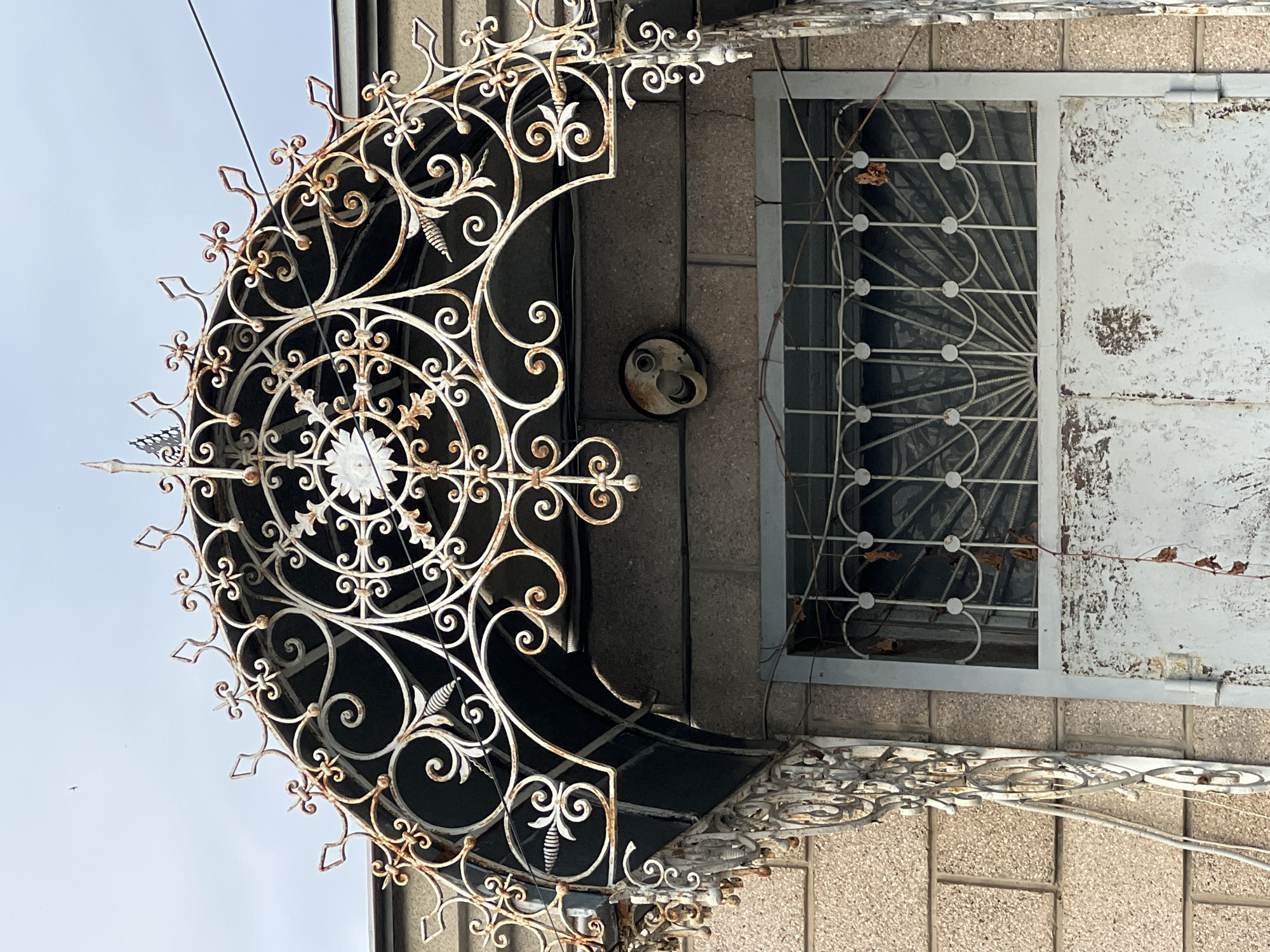 This image captures a close-up view of ornate, weathered white wrought ironwork above a textured, grey stone-tiled facade. The ironwork, likely part of a balcony or an awning, features intricate scrollwork, floral motifs, and pointed decorative elements. Some sections show signs of rust, suggesting age and exposure to the elements.
To the right of the ironwork, a window is visible with a similarly decorative metal grate. The window itself appears to be dirty, and dried leaves are caught within the grate, hinting at an outdoor setting. Beside the window, a section of the building wall is painted white, with significant chipping and peeling, revealing darker layers underneath. A light fixture, also white and circular with exposed wiring, is mounted on the wall between the ironwork and the window.
The overall impression is one of an aged, perhaps historical, building exterior. The lighting suggests daytime, with a bright sky visible in the upper left quadrant. No people are present in the frame. The setting appears to be outdoors, possibly a residential or commercial building in Krasnodar, Russia, given the location context. There is no visible text in the image.