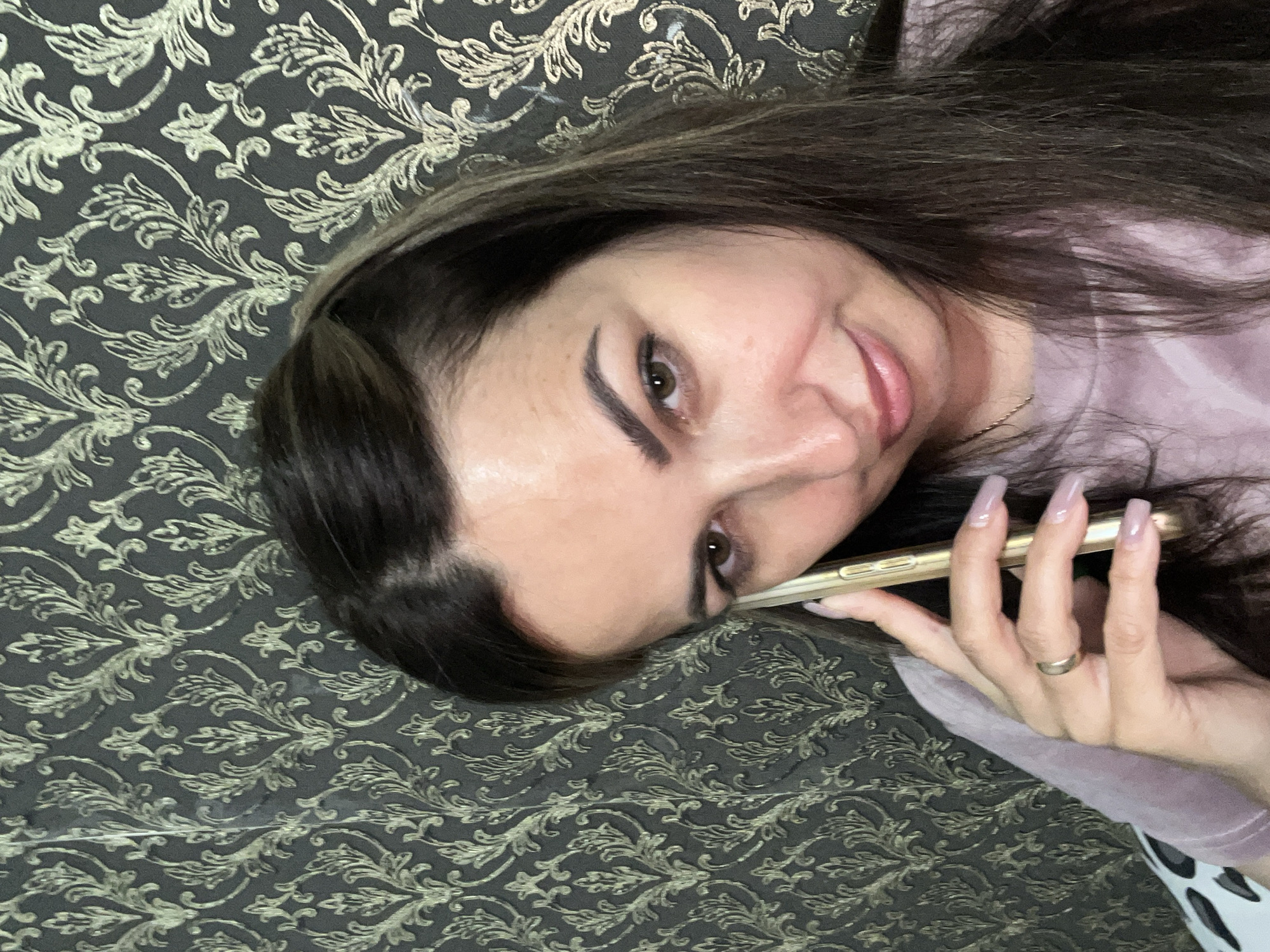 A medium shot shows a woman holding a gold smartphone to her right ear. She has long, dark brown hair that is partially pulled back. Her skin is light-toned with some freckles on her cheeks and nose. Her eyes are brown and she is looking towards the camera with a slight smile. She is wearing a light purple long-sleeved shirt. A ring is visible on her left ring finger. The background is a dark grey wallpaper with an ornate gold floral pattern. The scene appears to be indoors, possibly in a home.