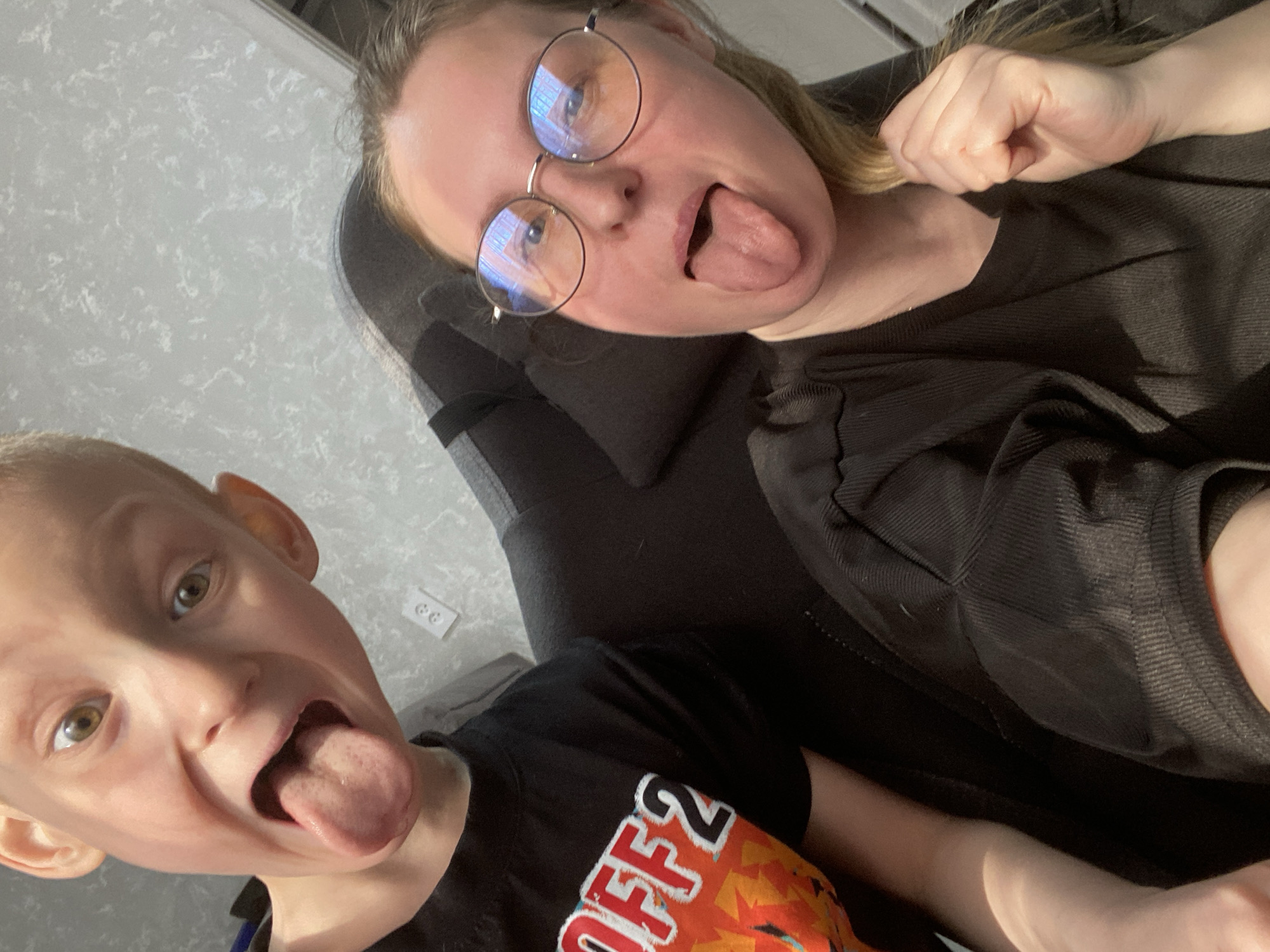 An indoor selfie captures a playful moment between two fair-skinned individuals, an adult female and a young male child, both sticking out their tongues. The adult female, with light hair and wearing round-rimmed glasses and a black top, is positioned in the upper right, seated on a dark chair or sofa. Reflections of indoor light sources are visible in her glasses. The young child, with light hair, is in the lower left, wearing a black t-shirt that features a graphic with the visible text "OFF2" and an orange, flame-like pattern below it.
The setting appears to be a home in Noviy, Russia, characterized by a light gray, textured wall in the background. An electrical outlet with two receptacles is visible on the wall behind the child. The lighting is bright, suggesting it is daytime. The overall mood is informal and jovial, highlighting a moment of lighthearted interaction.